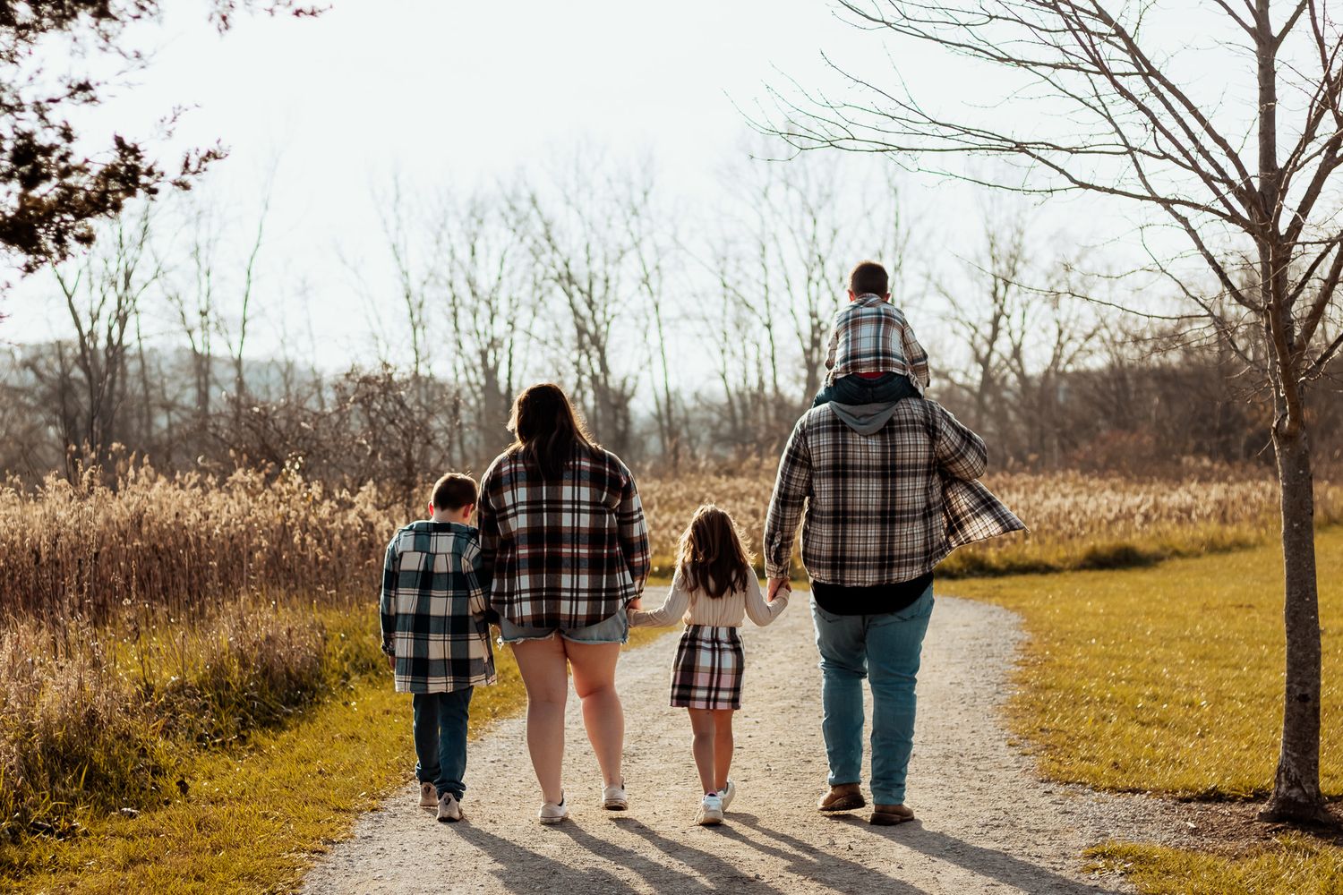 Family wearing matching plaid shirts walk together on a nature trail during autumn with tall grass and trees.