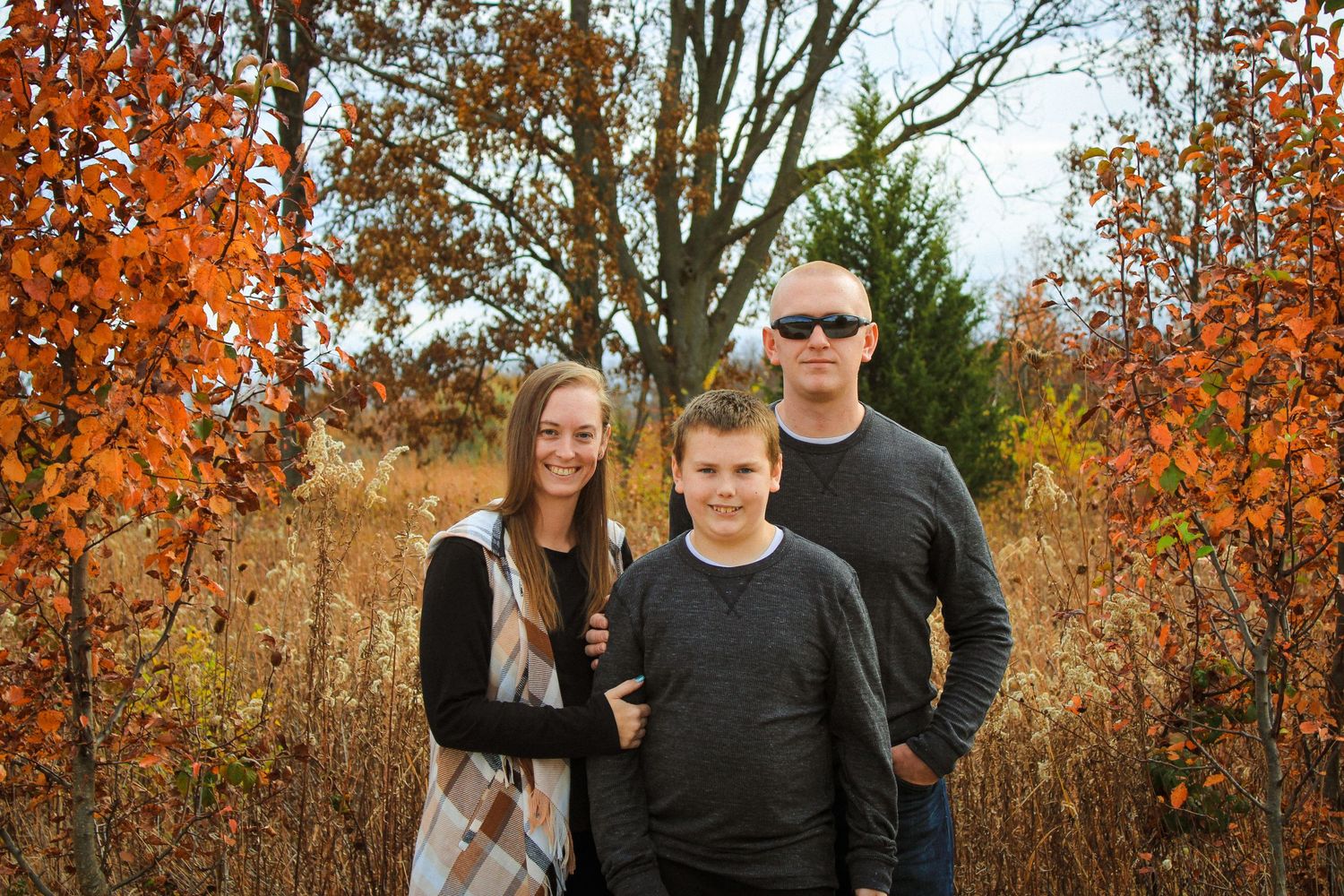 Outdoor autumn portrait of three people standing together surrounded by orange fall foliage.