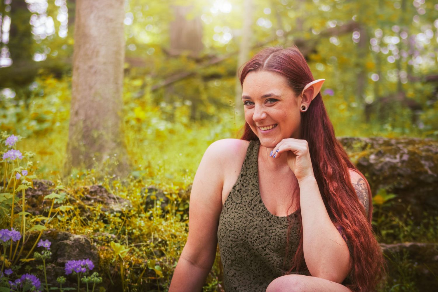 Long-haired person in a green tank top smiles in a sunlit forest setting with moss and wildflowers in the background.