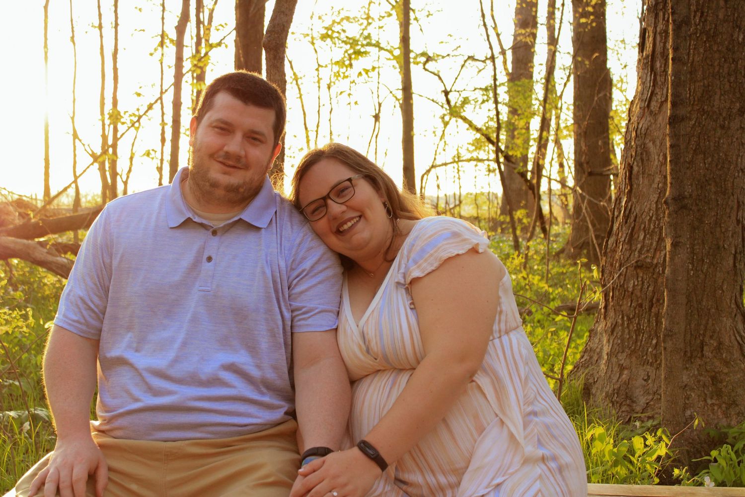 A couple sits together on the ground in a sunlit forest during golden hour.