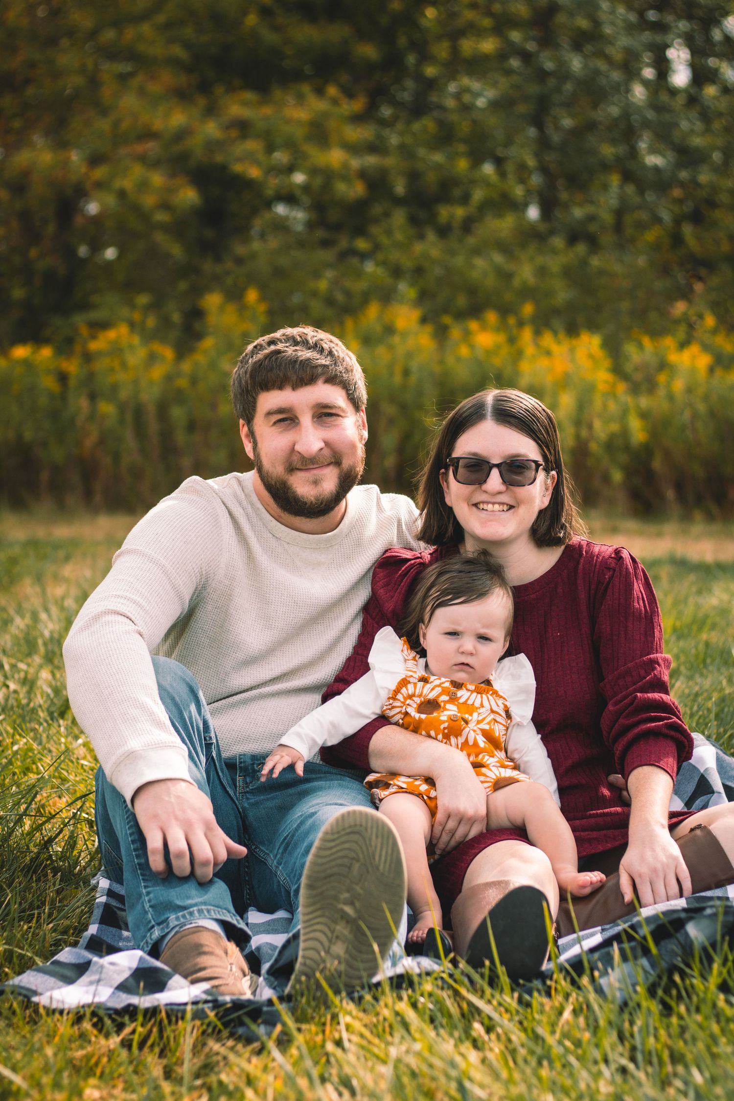 A family sits together on a blanket in an autumn field with golden wildflowers and trees in the background.