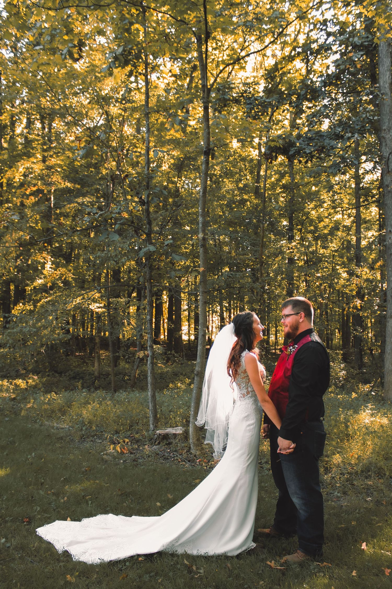 A couple in wedding attire stand together in a sun-dappled forest with golden autumn leaves and flowing white veil.