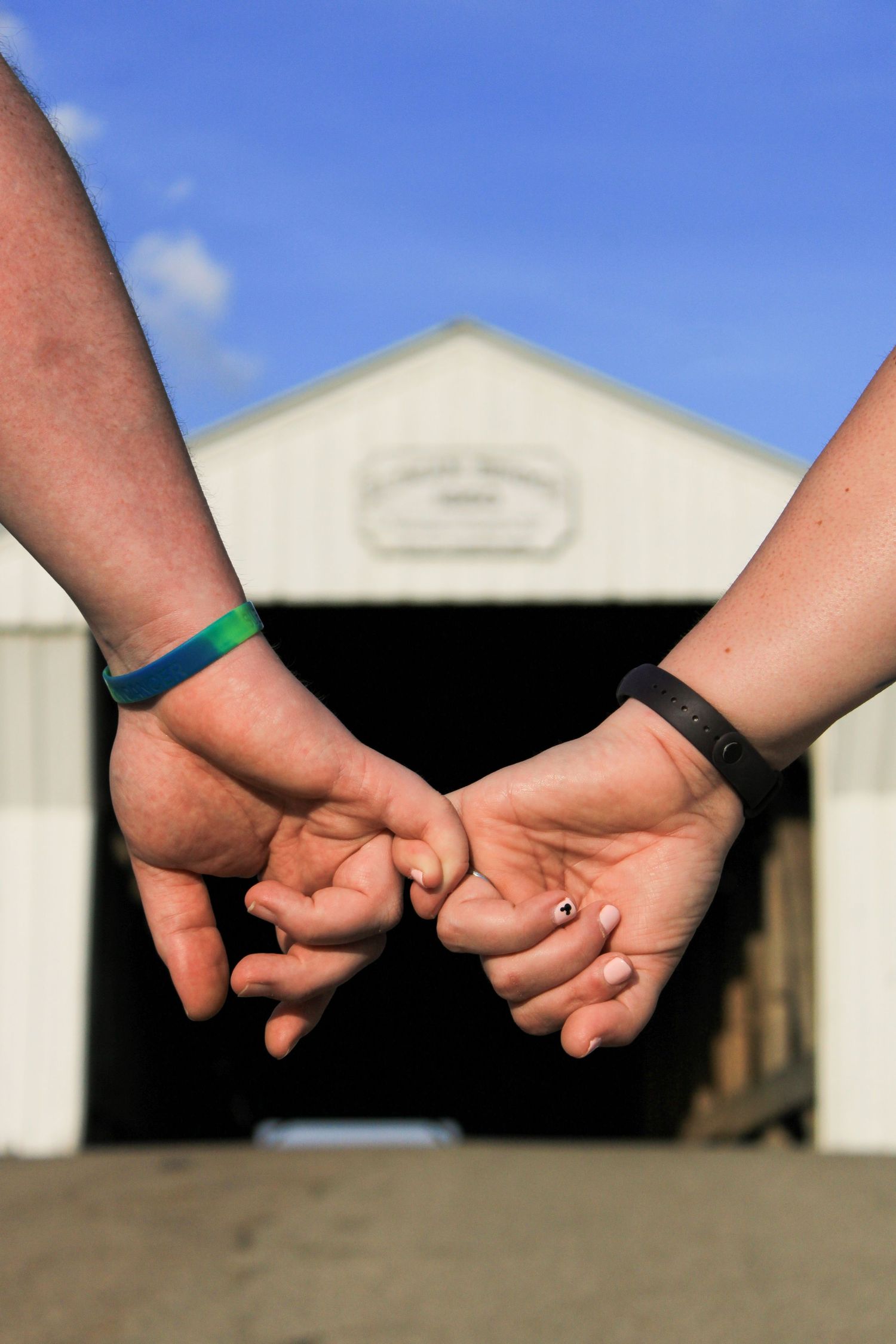 Two hands wearing fitness bands reach behind to hold pinkies together in front of a white barn style bridge.