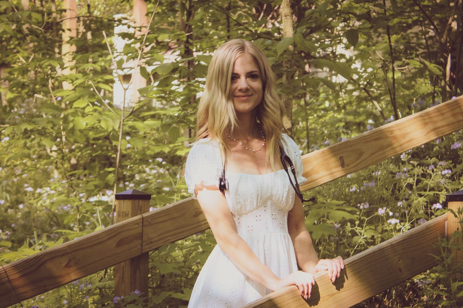 A person in a white summer dress poses on a rustic wooden bridge surrounded by lush greenery in warm sunlight.