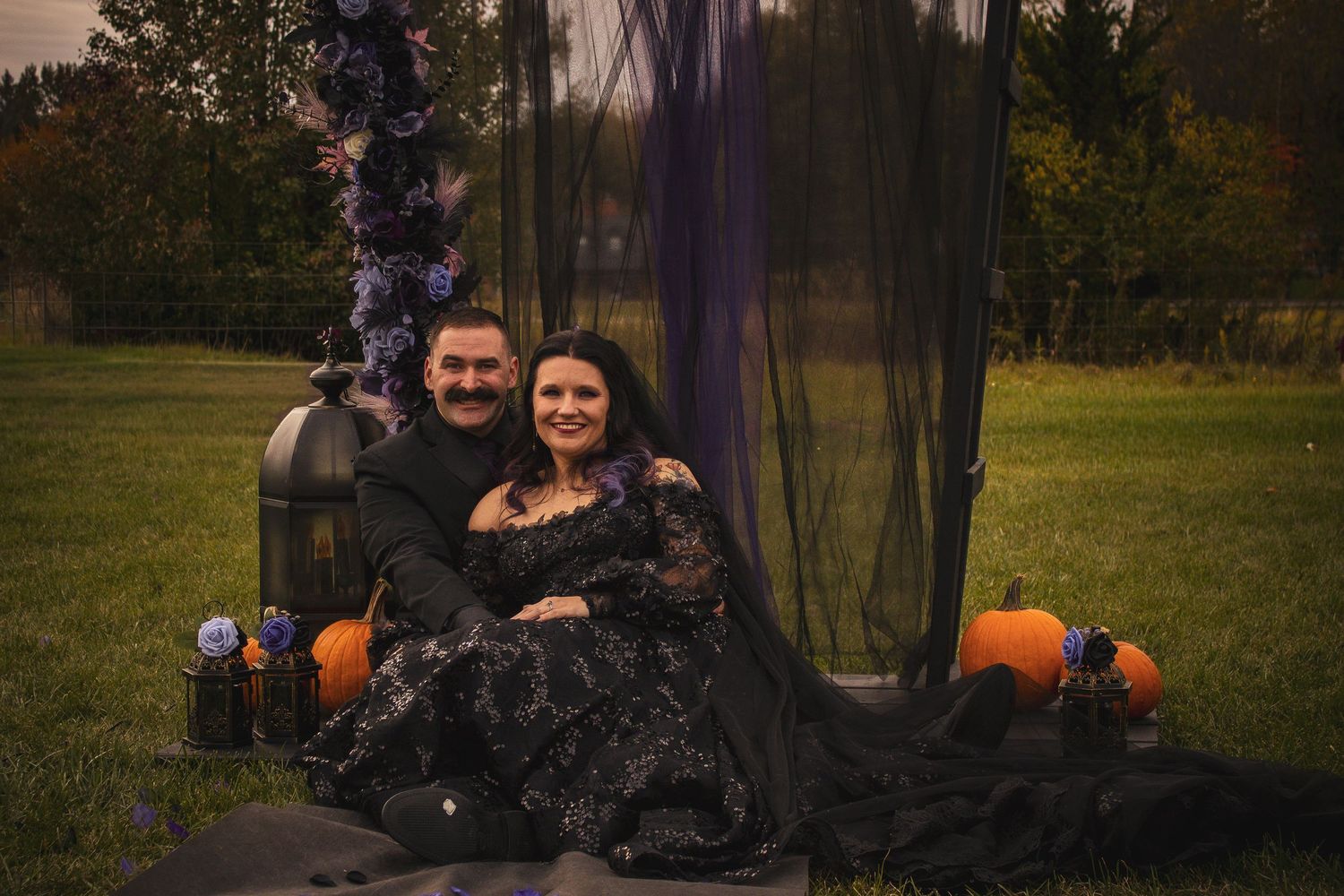 A couple sits together on a blanket surrounded by Halloween decorations including pumpkins and black tulle fabric outdoors.