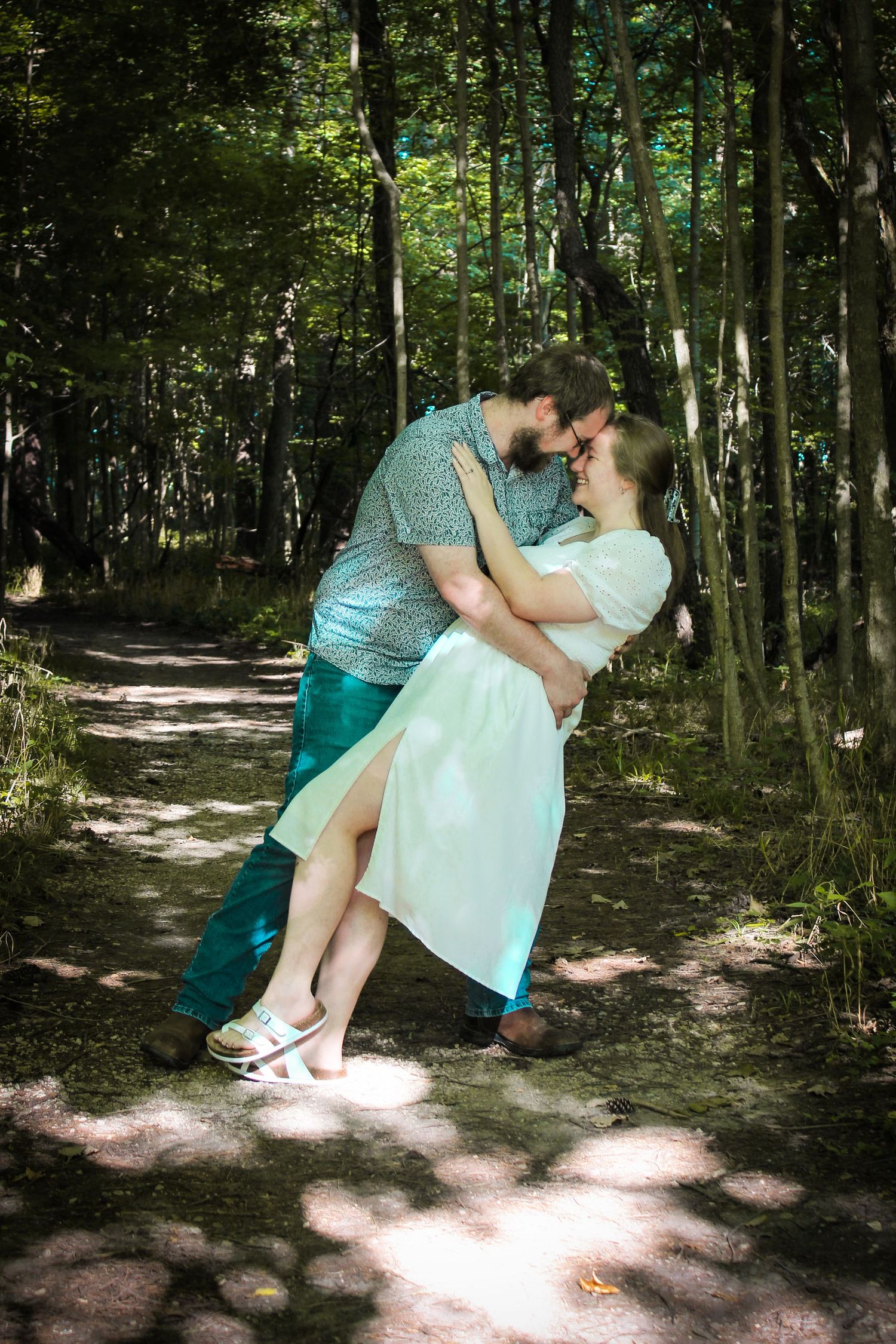 A romantic couple shares a tender moment on a sunlit forest path surrounded by green trees.