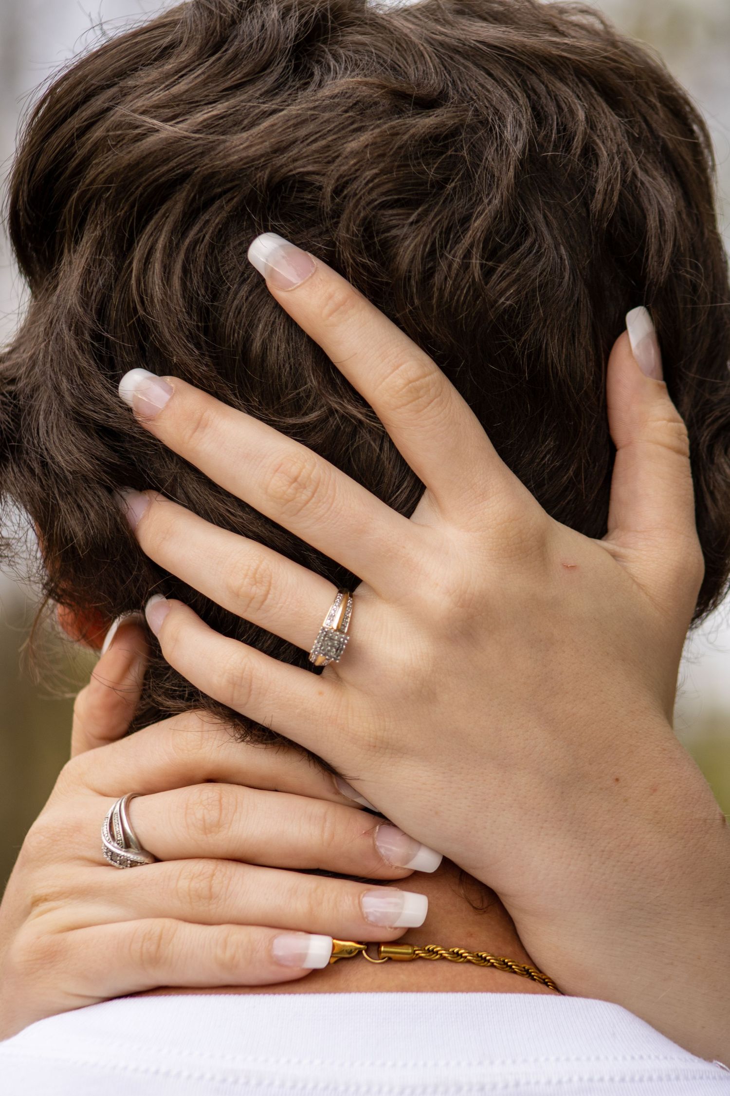 Hands wearing silver an engagement ring intertwined in dark brown hair against a blurred outdoor background.