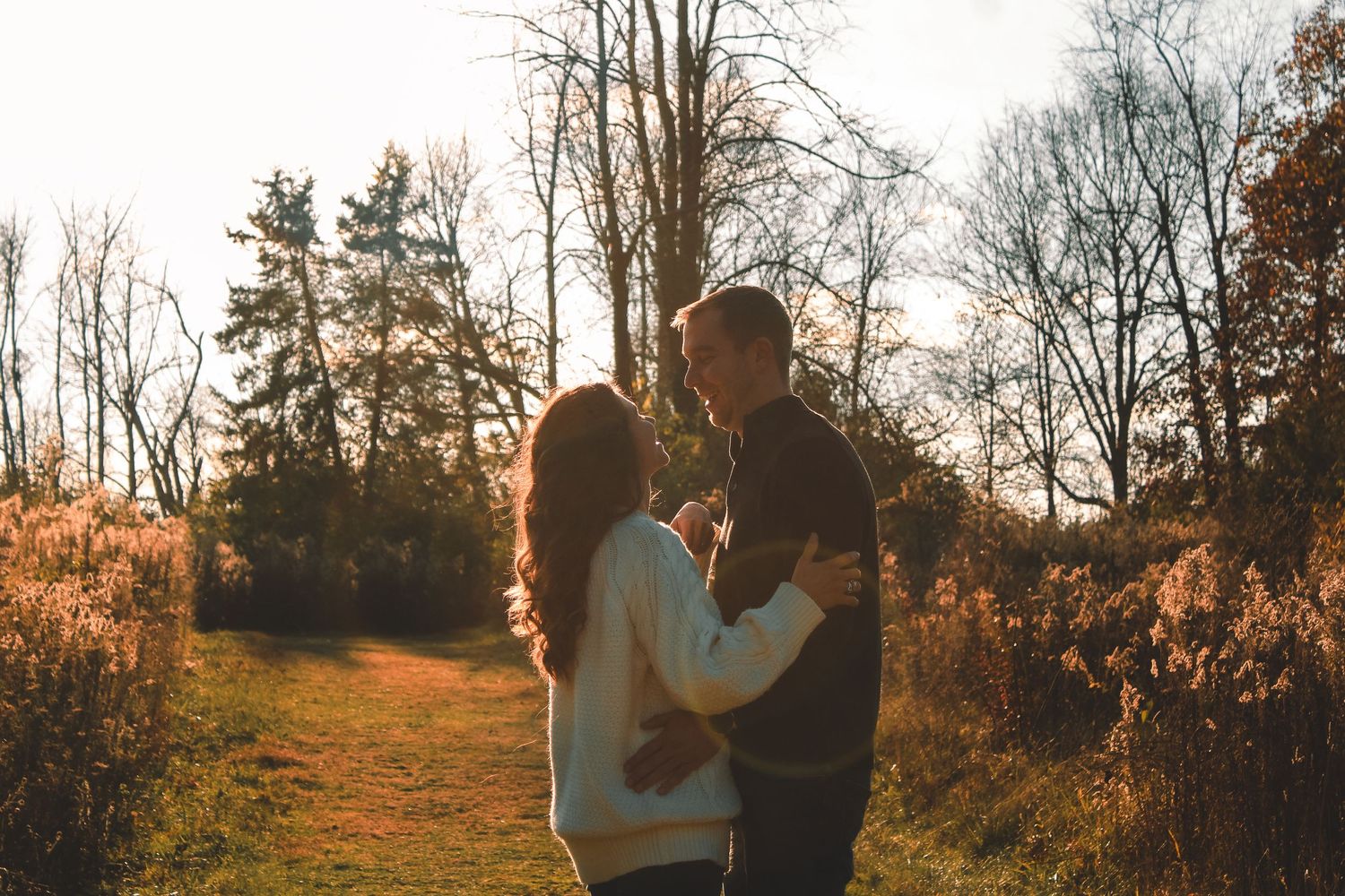 A couple embraces in golden sunlight on a wooded trail during autumn sunset creating a warm silhouette against nature.