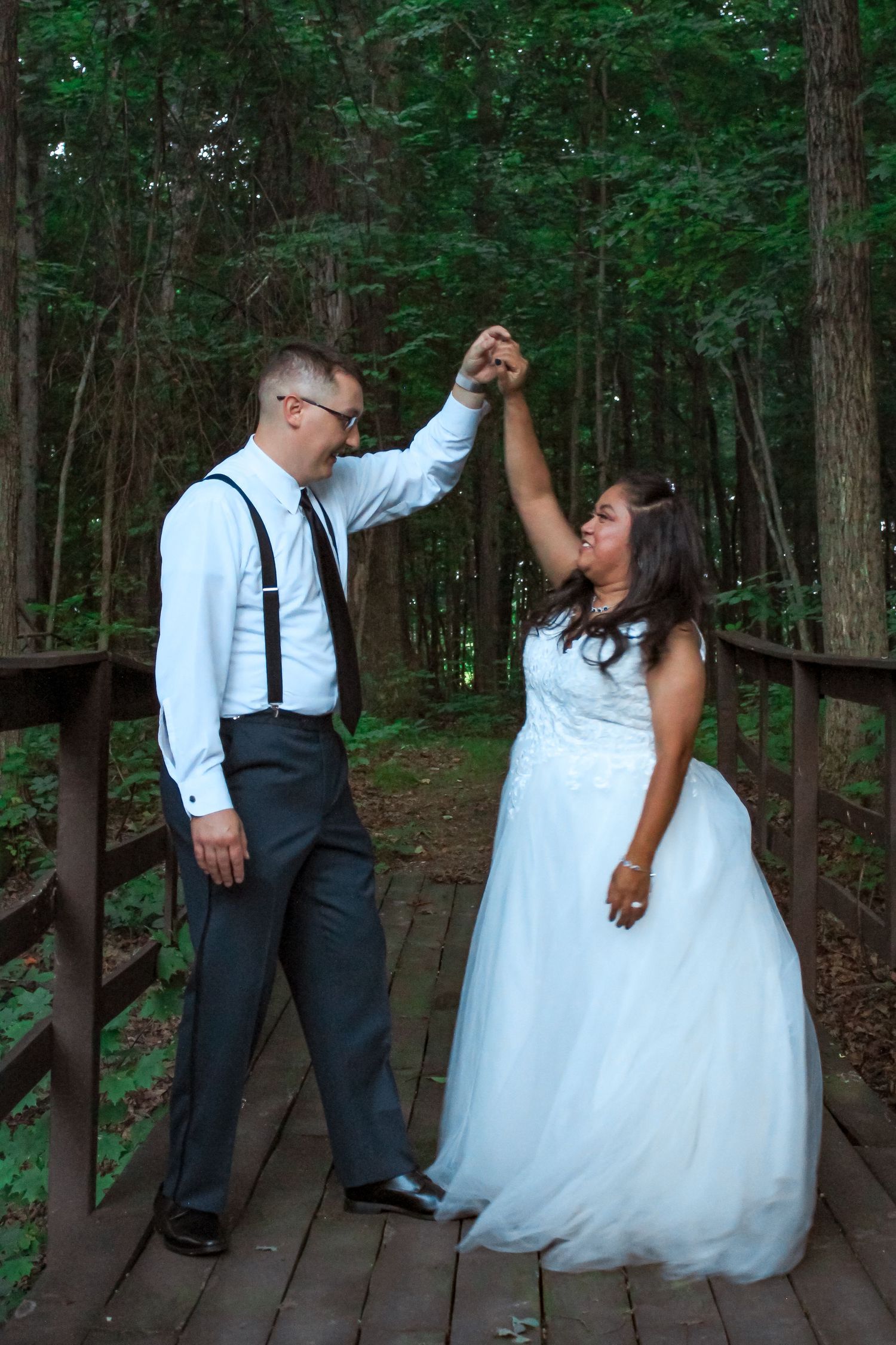 A couple in wedding attire dancing together on a wooden deck surrounded by lush green forest.
