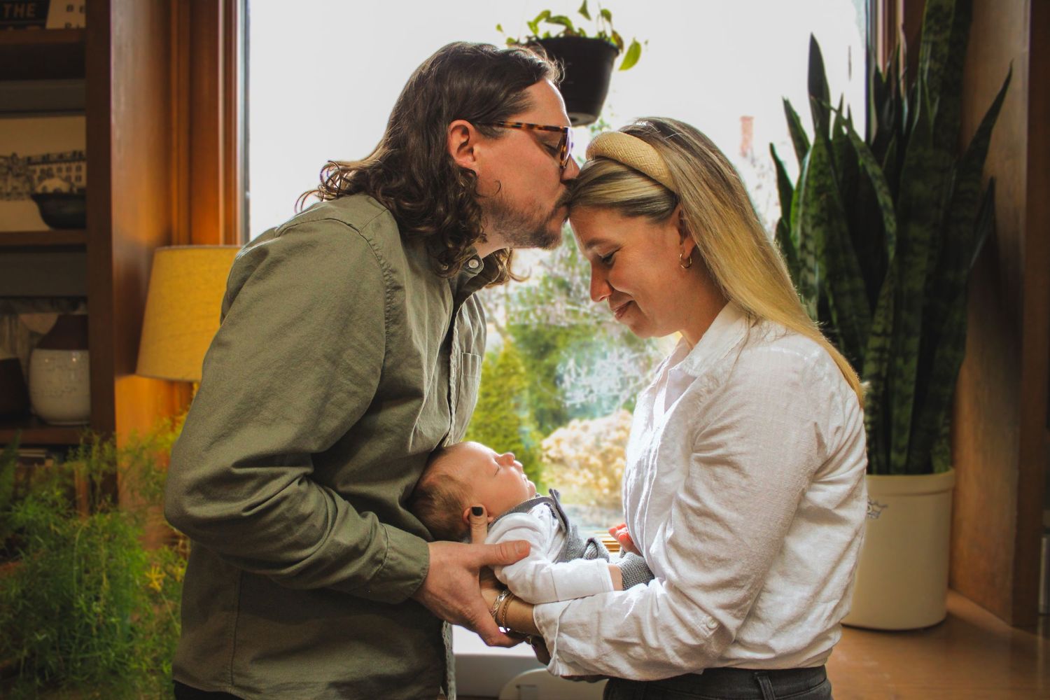 Two people share an intimate moment while sitting together indoors near a window with houseplants in the background.
