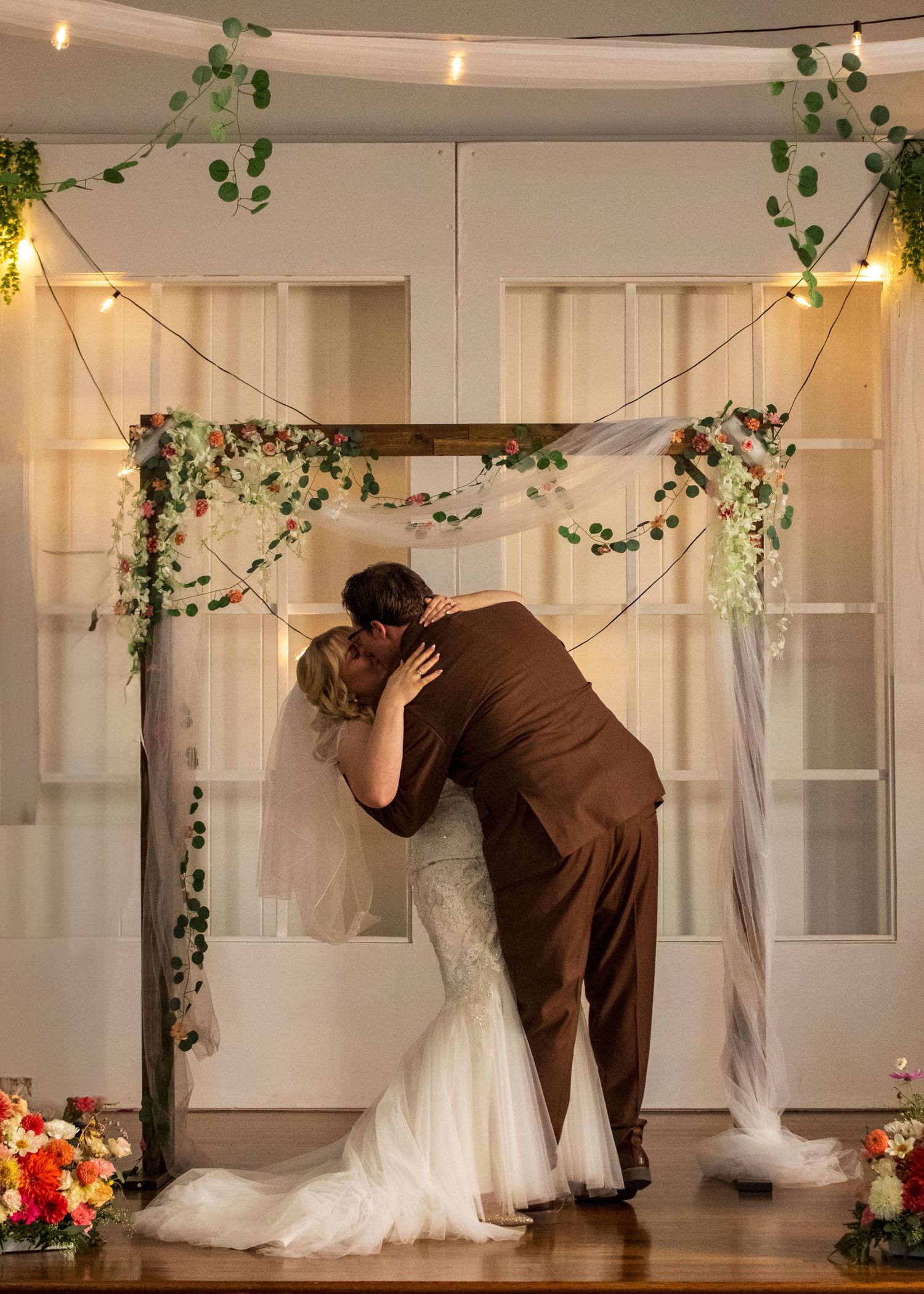 Bride and groom share first kiss under floral arch with string lights at indoor wedding ceremony.