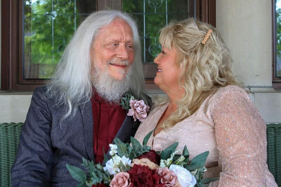 Elderly couple smiling at each other during wedding ceremony, man with long white hair and beard, woman in pink dress holding bouquet.