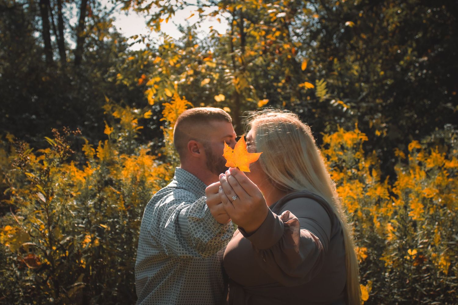 A couple hiding behind a leaf together in a field of golden wildflowers at sunset with warm sunlight glowing through the trees.
