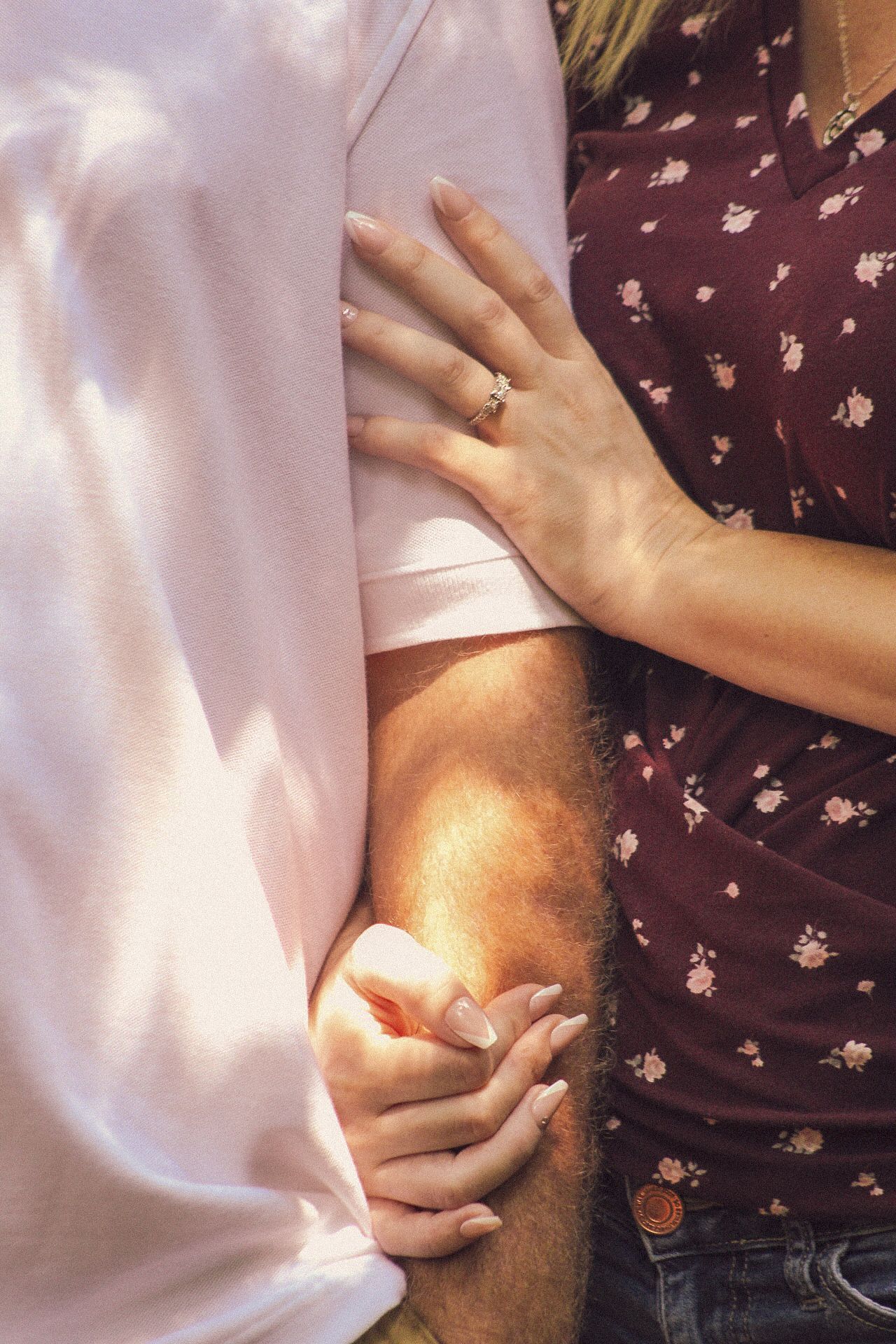 Close-up of hands showing an engagement ring with dappled sunlight on a purple floral dress and pink shirt.