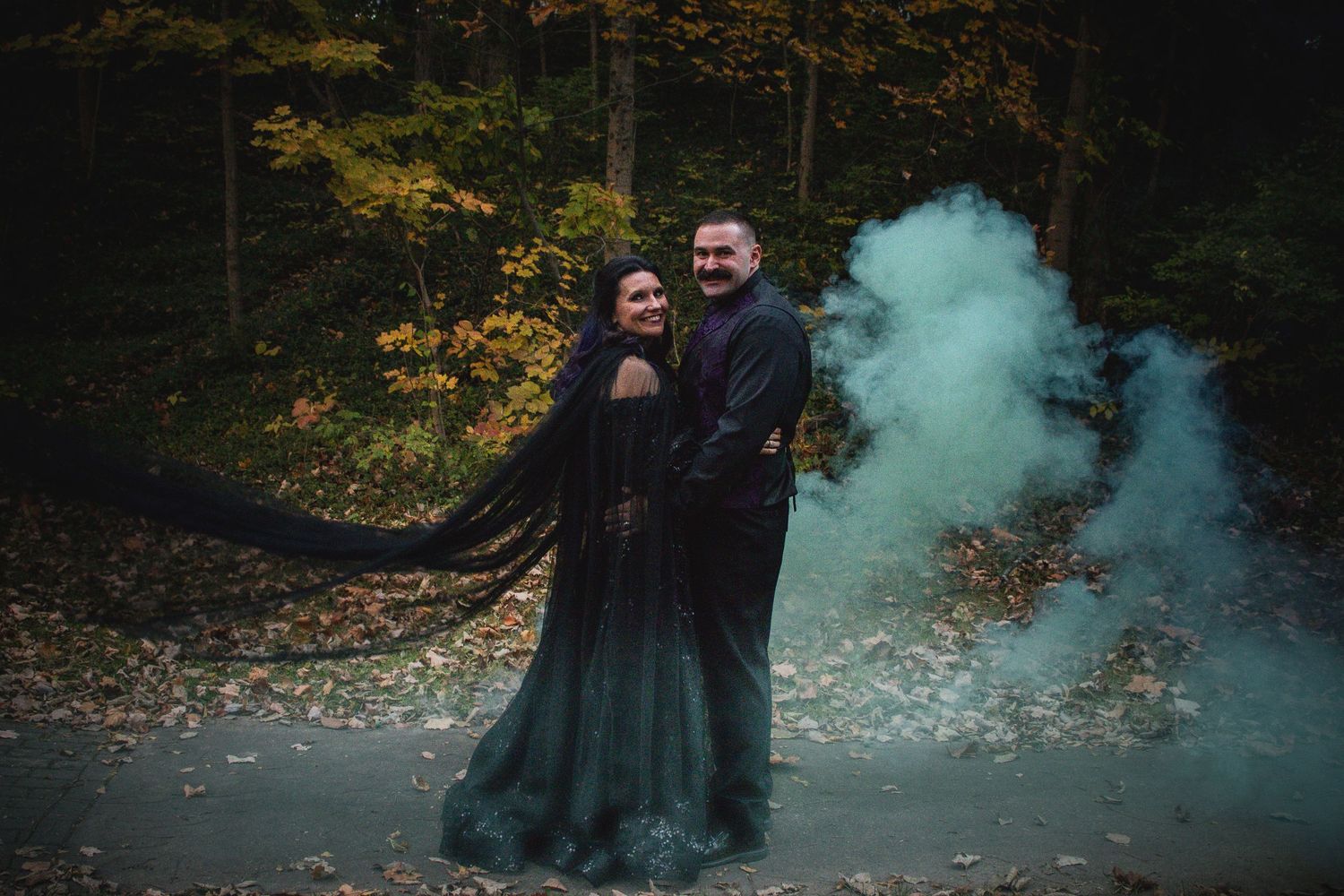 A couple stands together surrounded by moody teal smoke in an autumn forest setting.