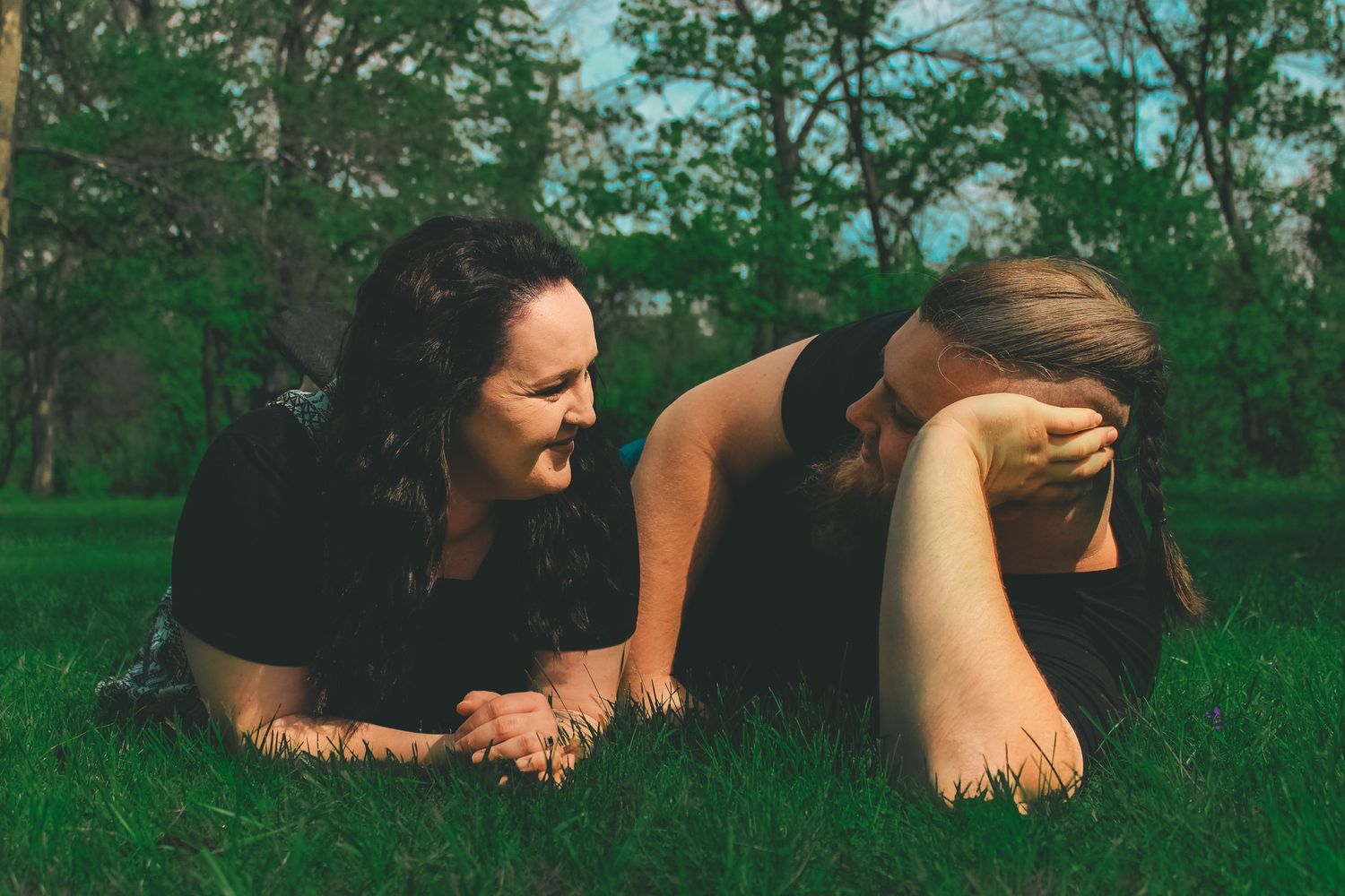 Two people laying in green grass on a sunny day under leafy trees.
