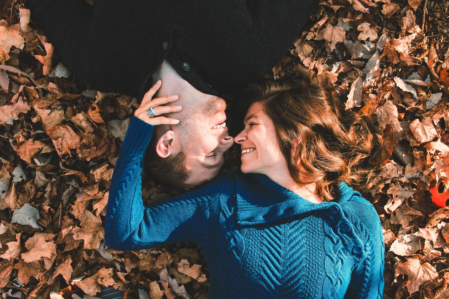 A couple in black and blue sweaters lying on autumn leaves while sharing an intimate moment.