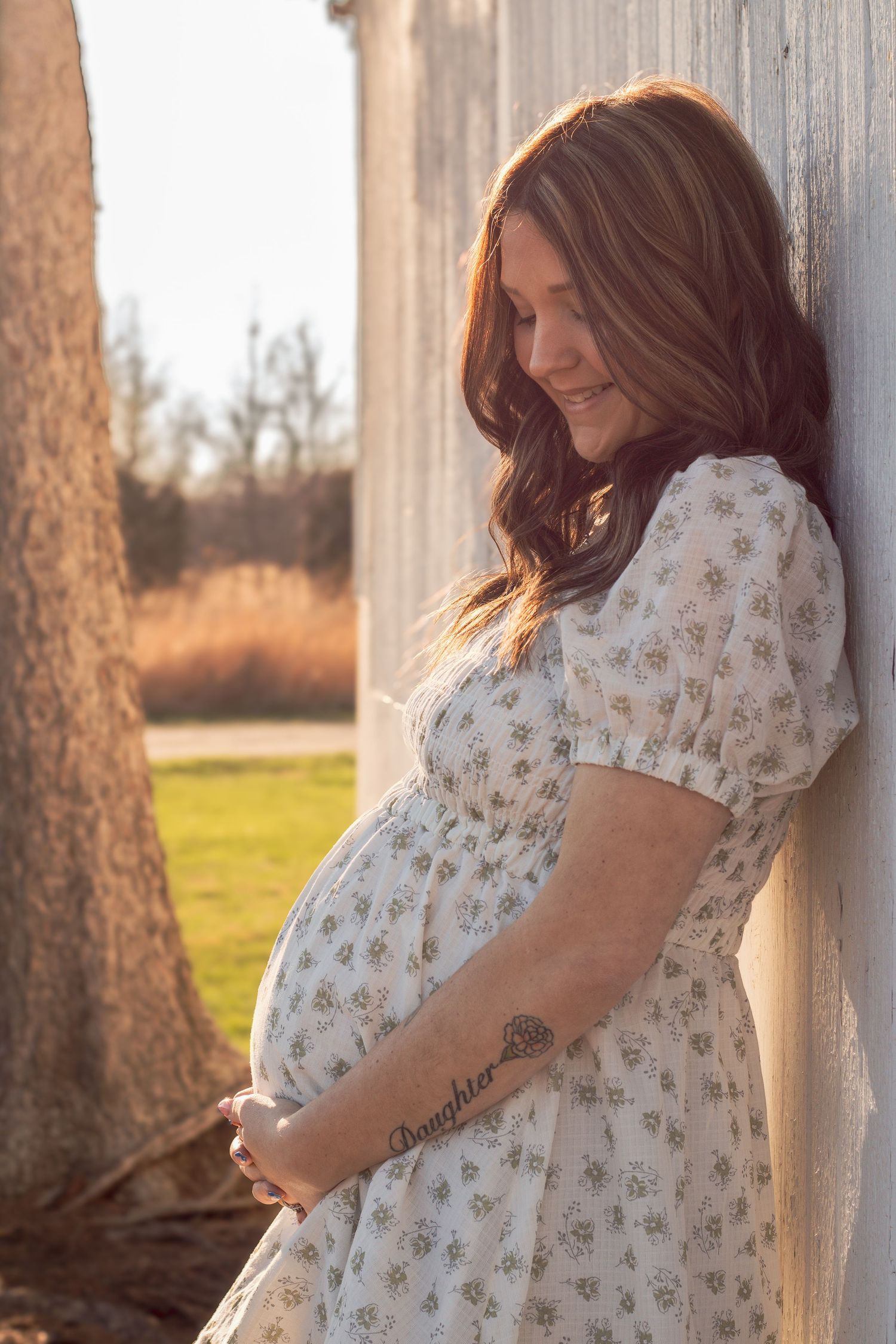 Pregnant woman in floral dress cradling baby bump outdoors, leaning against rustic wall in golden hour light.