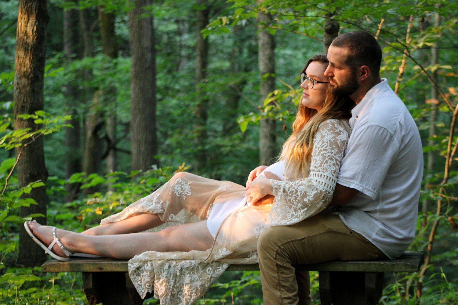 A romantic couple sits together on a rock in a lush green forest during an outdoor photoshoot.
