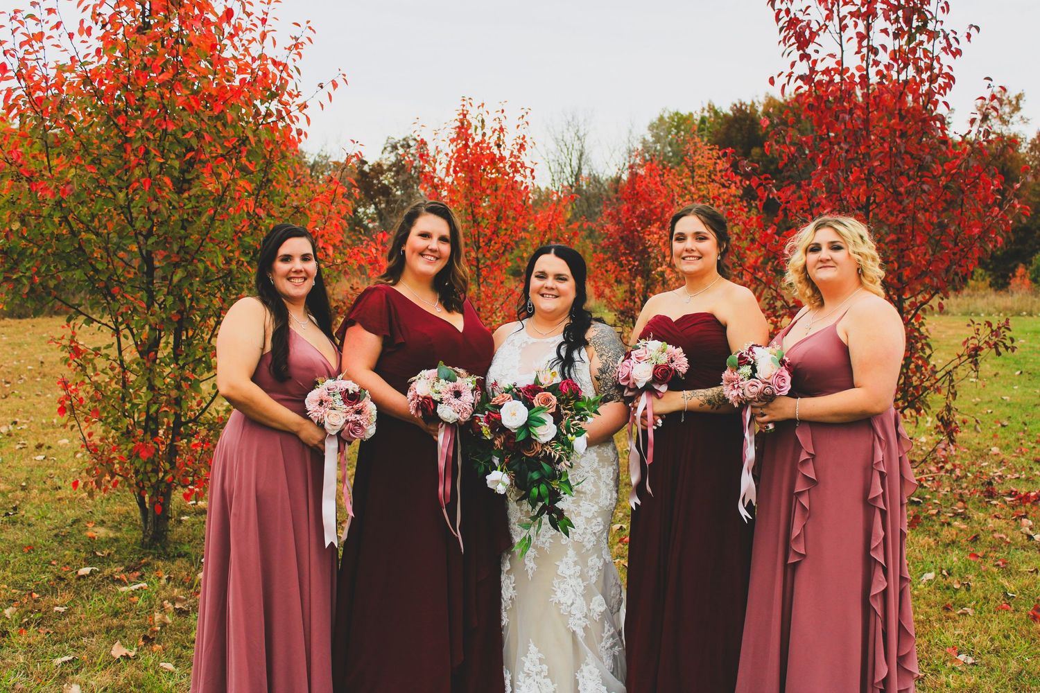 Bride in white lace gown poses with four bridesmaids in dusty rose dresses holding bouquets amid vibrant fall foliage.