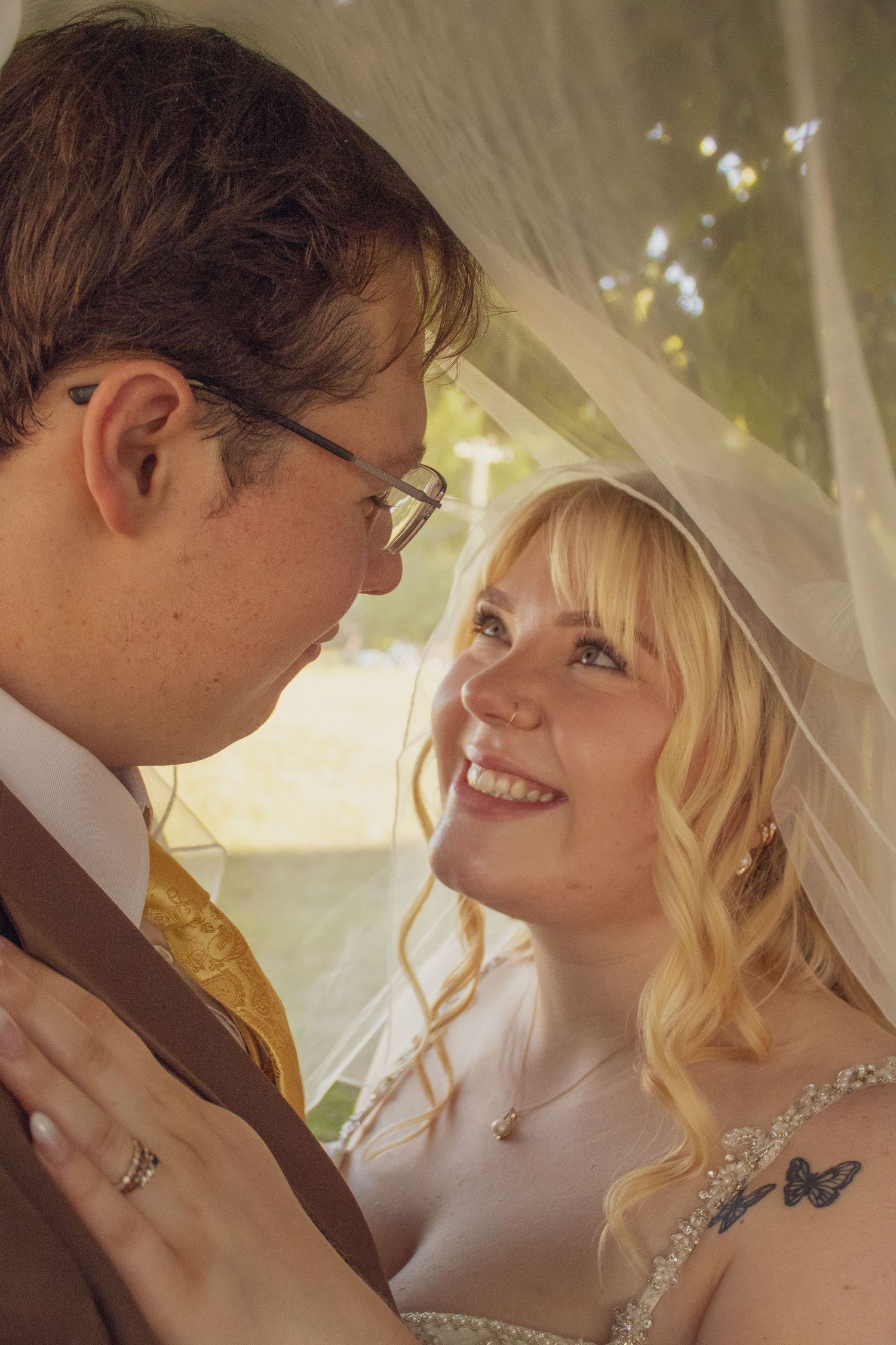 A smiling blonde bride in a beaded gown shares a romantic moment with her groom under a wedding veil outdoors.