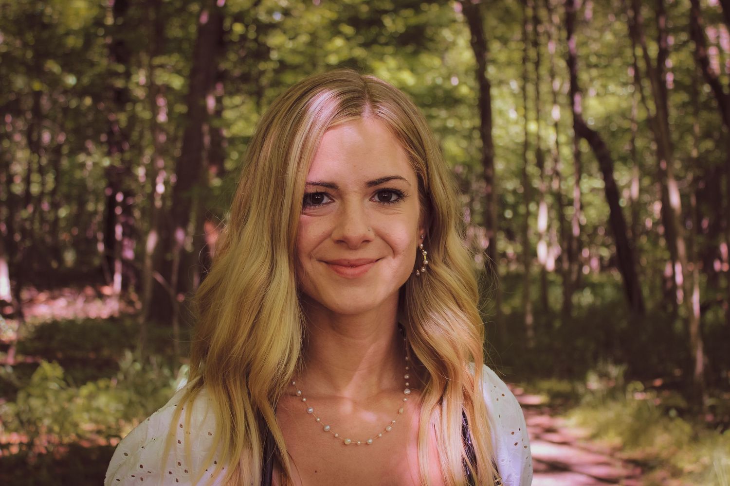 A smiling person in a light colored top poses outdoors in a wooded setting during golden hour.