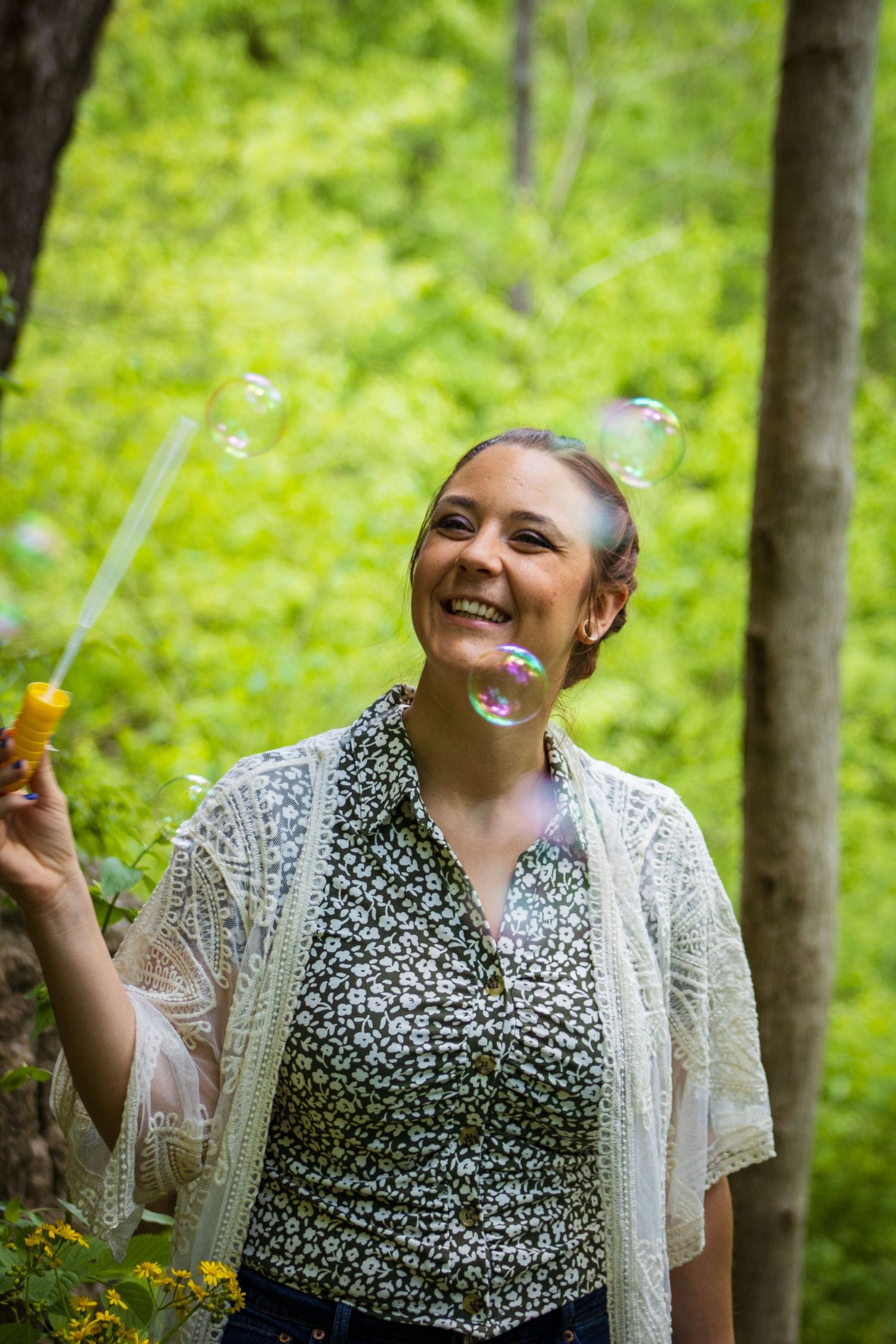 Someone blows bubbles in a forest setting while wearing a floral print top and lace cardigan.