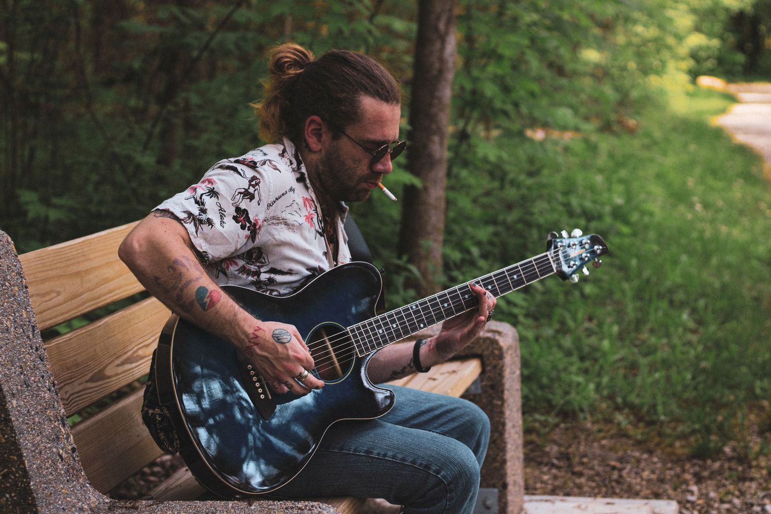 Musician with sunglasses and bun hairstyle plays electric guitar while sitting on wooden bench in wooded area.
