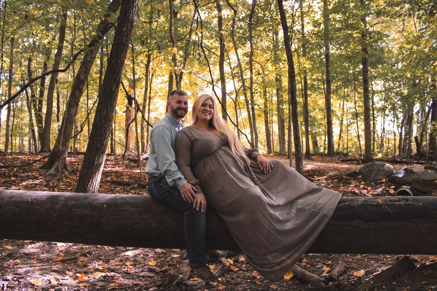 A couple sits together on a fallen tree log in an autumn forest during golden hour sunset.