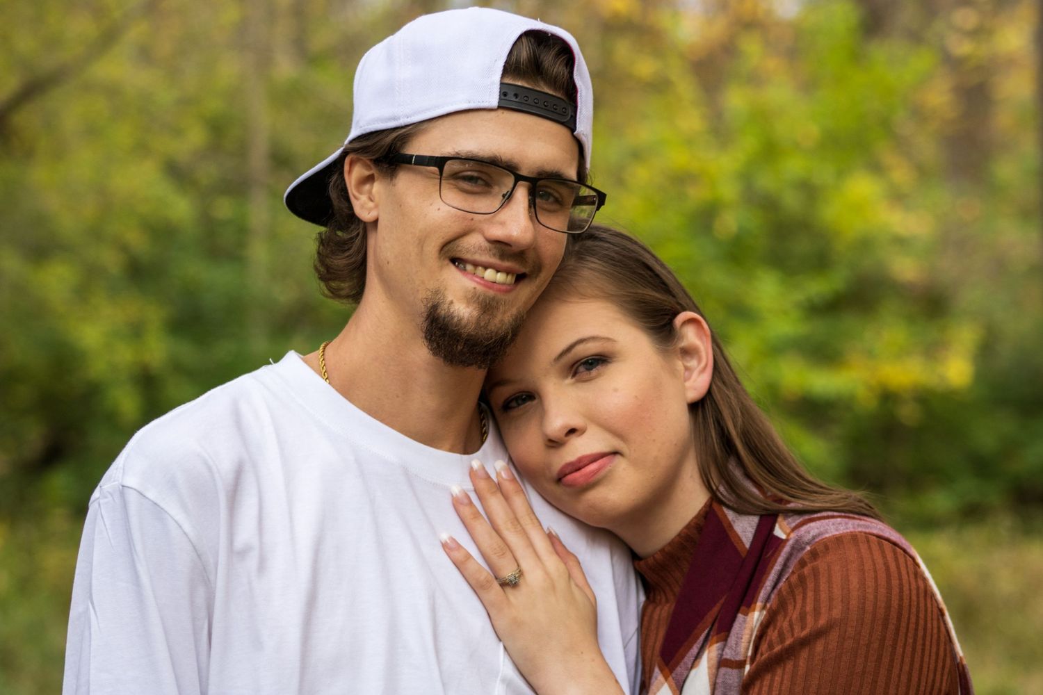 A couple wearing casual clothes embraces outdoors in a natural setting with soft green foliage in the background.