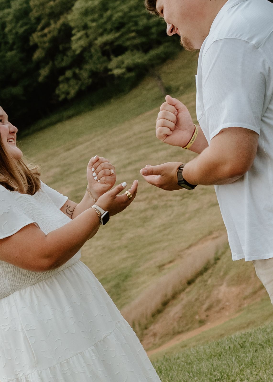Couple in white outfits playfully interact outdoors on a grassy hillside, laughing and reaching toward each other.