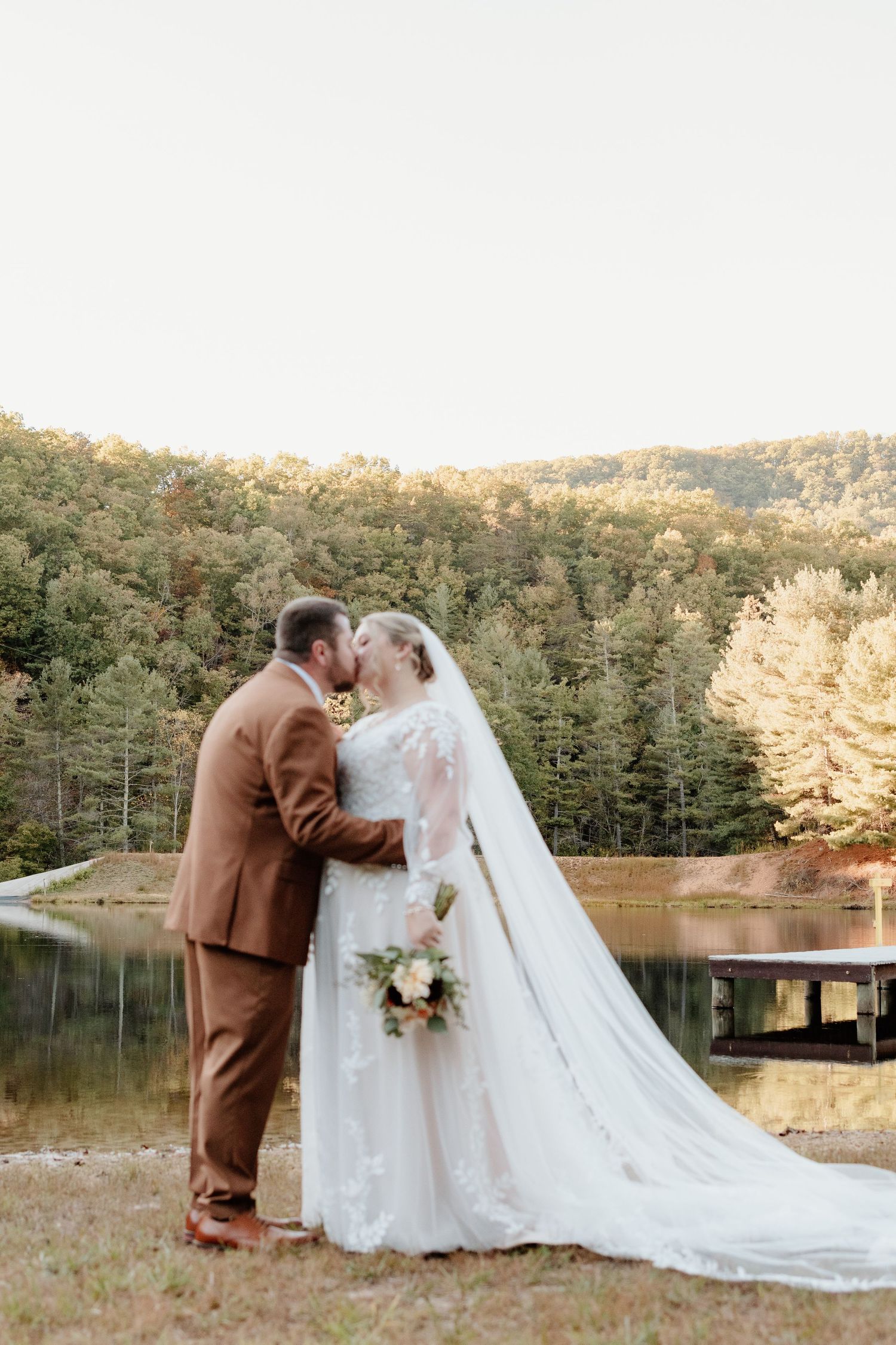 A couple shares an intimate moment by a lake during their autumn wedding ceremony with a scenic forest backdrop.