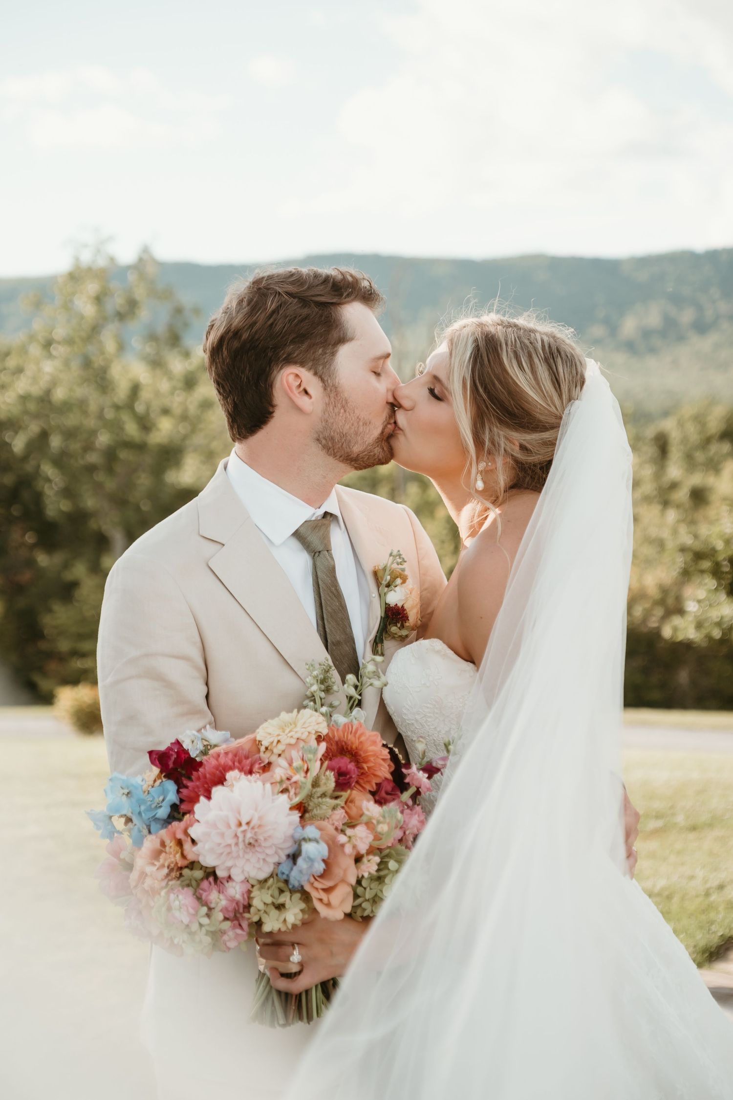 Bride and groom share a kiss outdoors, bride holds colorful bouquet with dahlias, wearing veil and strapless gown.