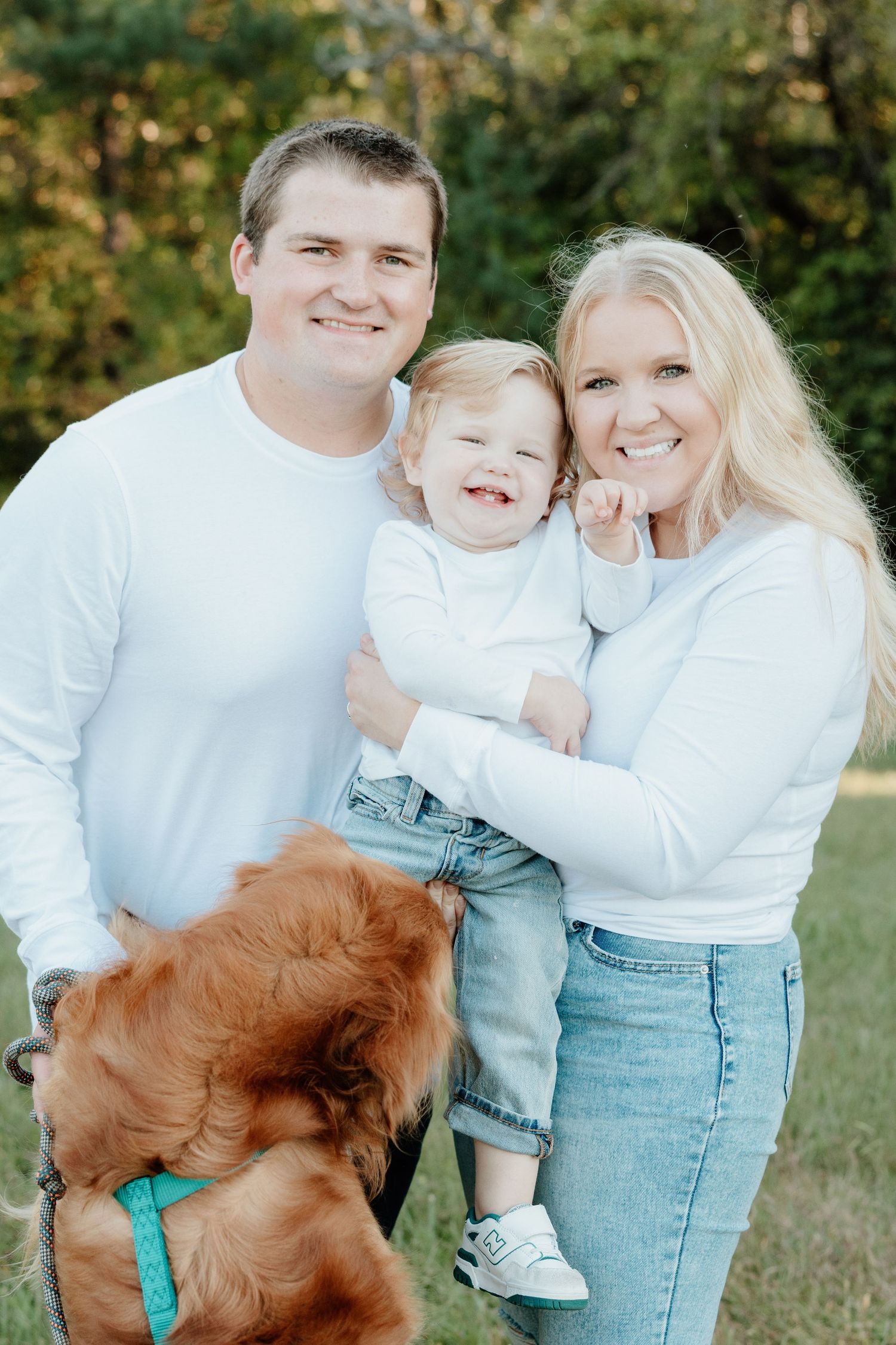 A family in matching white tops poses outdoors with their golden retriever dog wearing a teal collar.