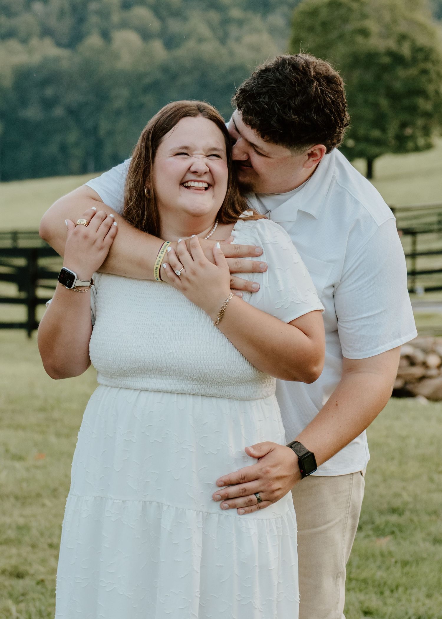 A couple embraces outdoors, woman in white dress laughing joyfully as man kisses her cheek on a green farm backdrop.