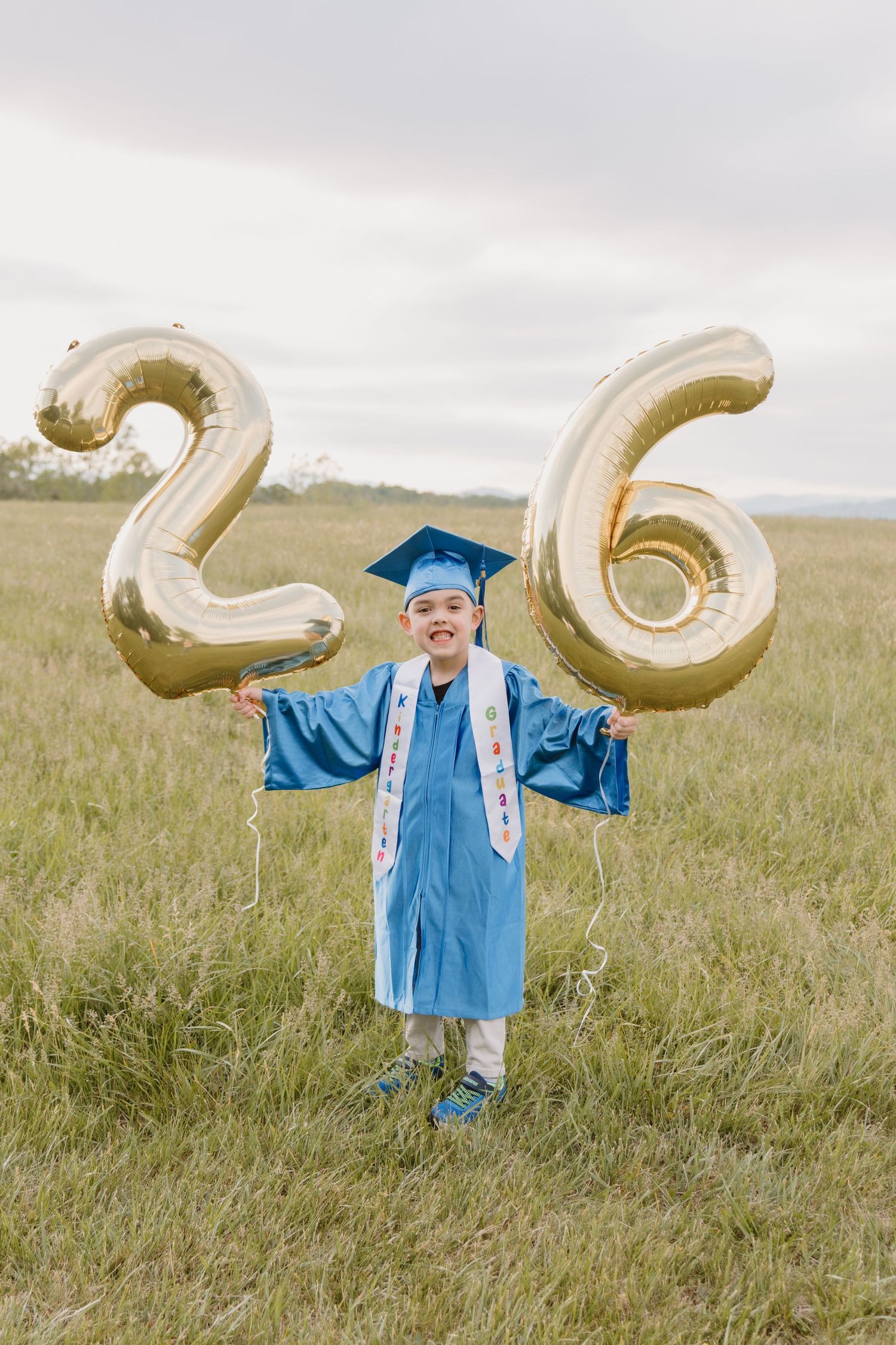 Child in blue graduation cap and gown holds gold number 26 balloons in a green field.
