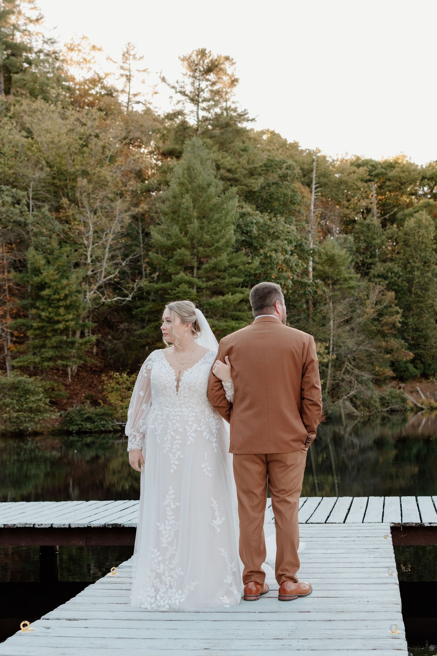 A couple shares a quiet moment together on a wooden dock overlooking a serene lake surrounded by autumn trees.