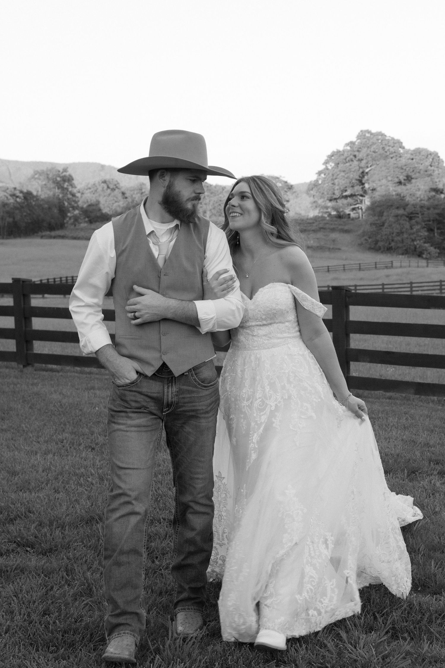 A couple in western wedding attire dances together in front of a rustic fence at sunset.