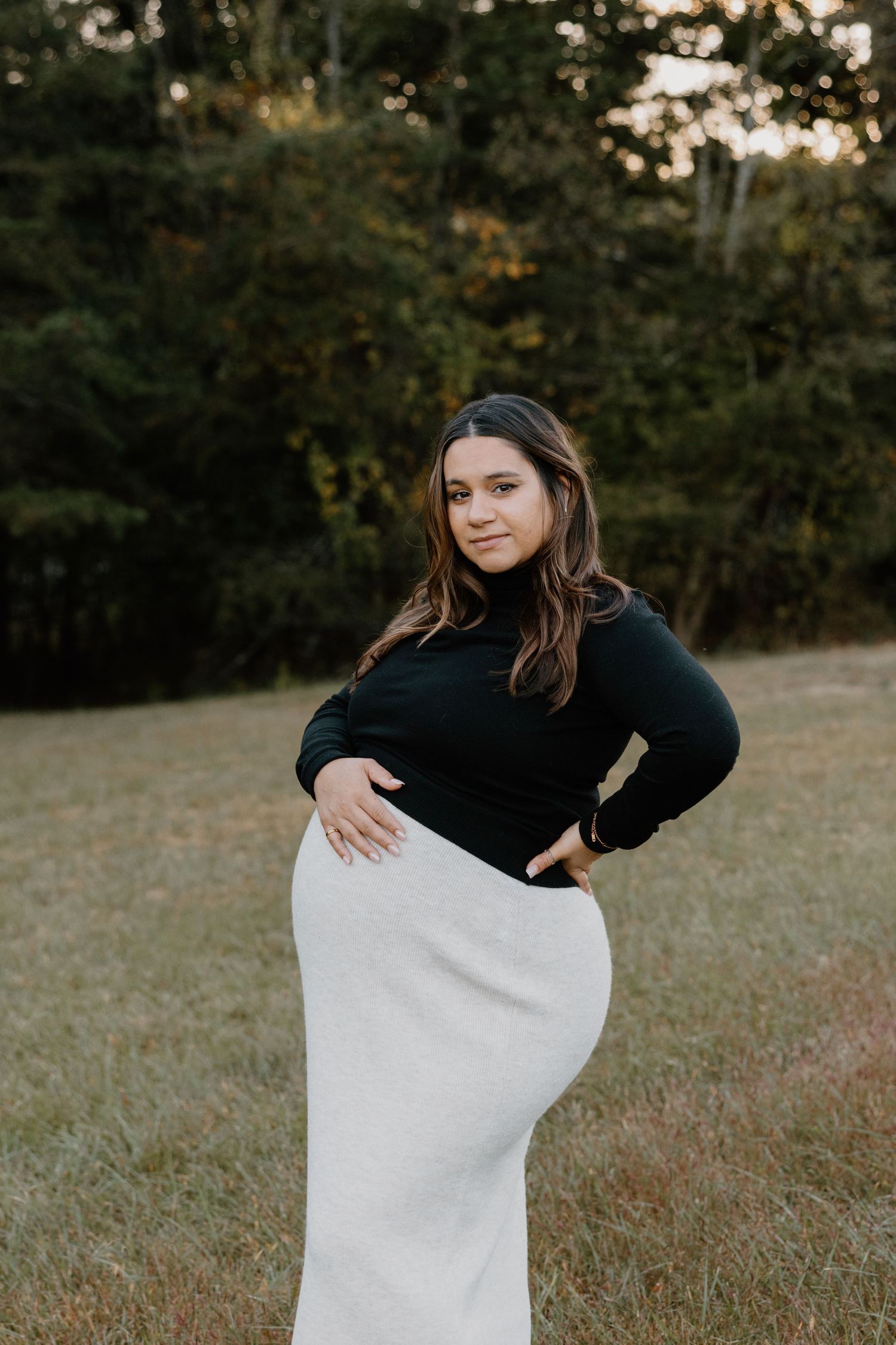 A woman in a black sweater poses in a grassy field during golden hour while holding a white blanket.
