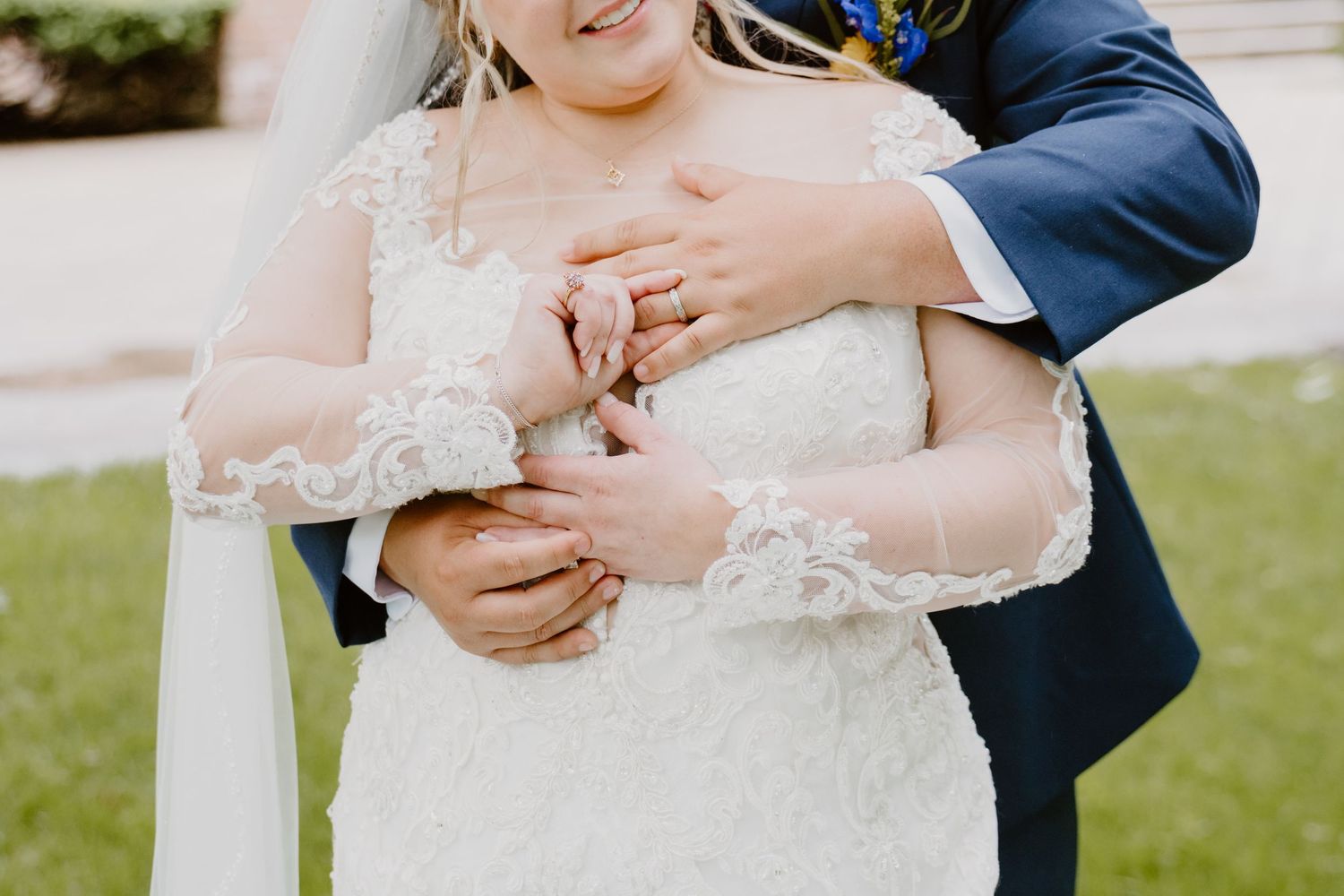 Bride in lace wedding gown embraced by groom in navy suit during outdoor wedding ceremony.