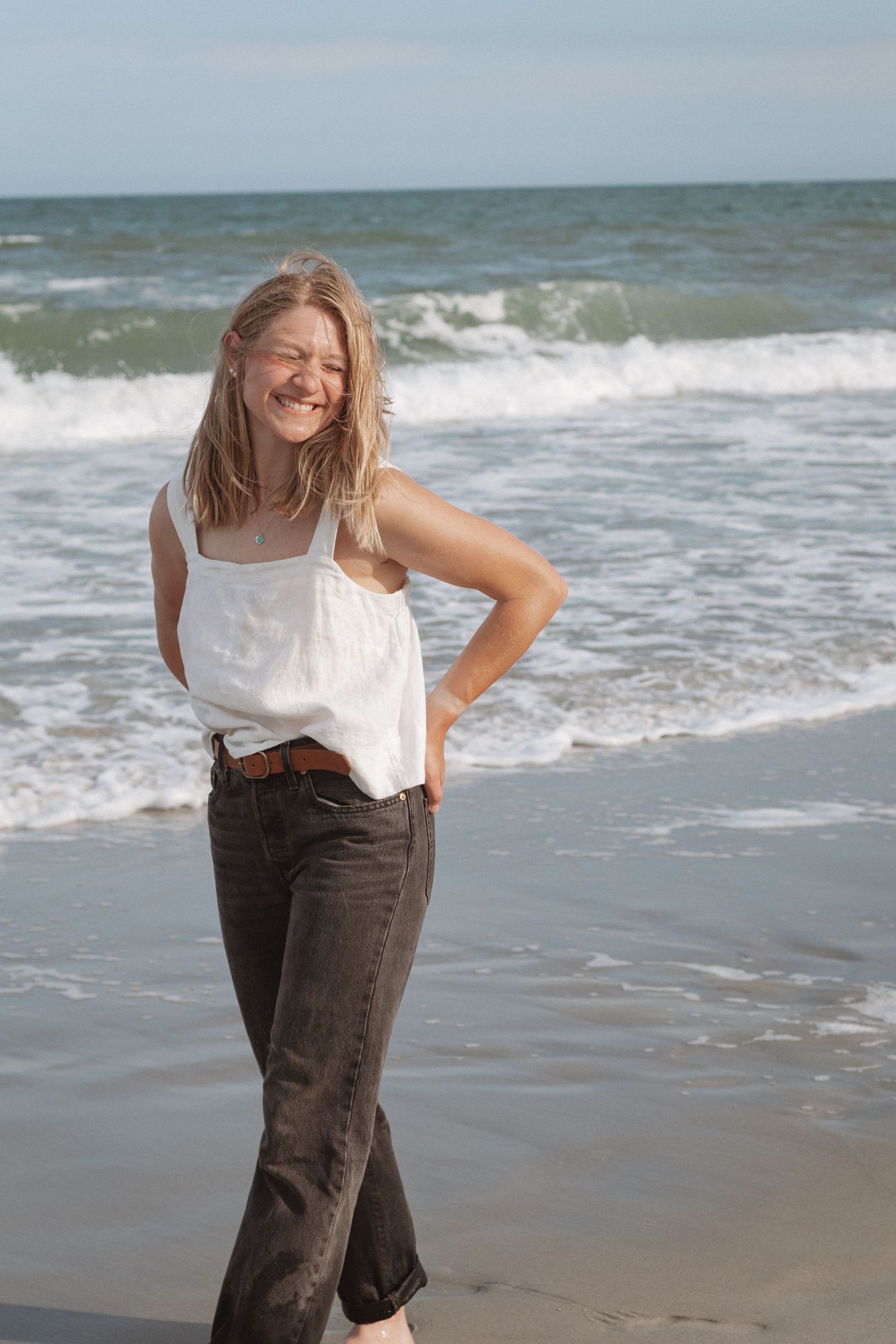 A person in a white top and brown pants poses joyfully on a sunny beach with crashing waves in the background.