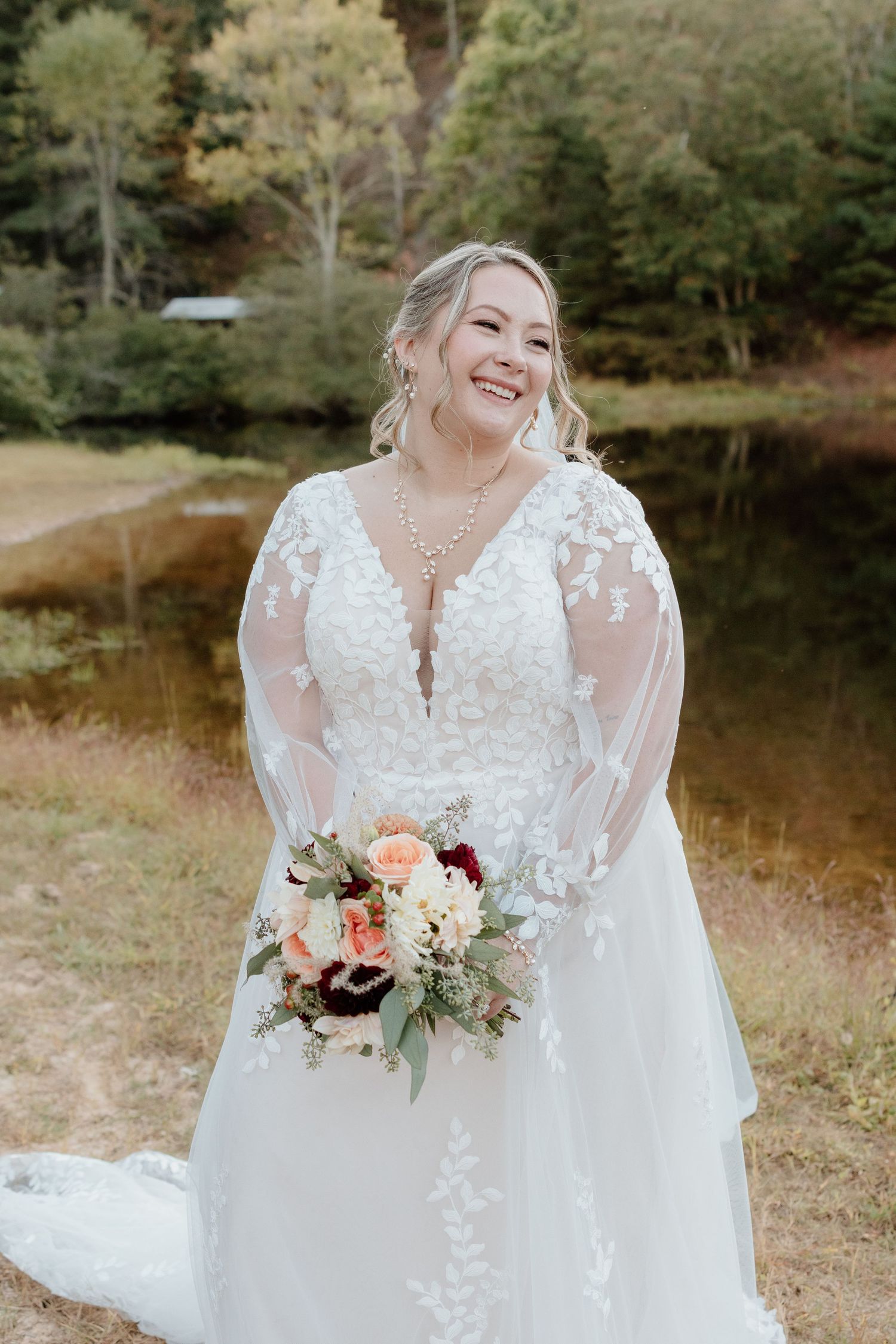 A bride in a lace long-sleeve wedding gown holds a peach and white bouquet by a serene pond in autumn.