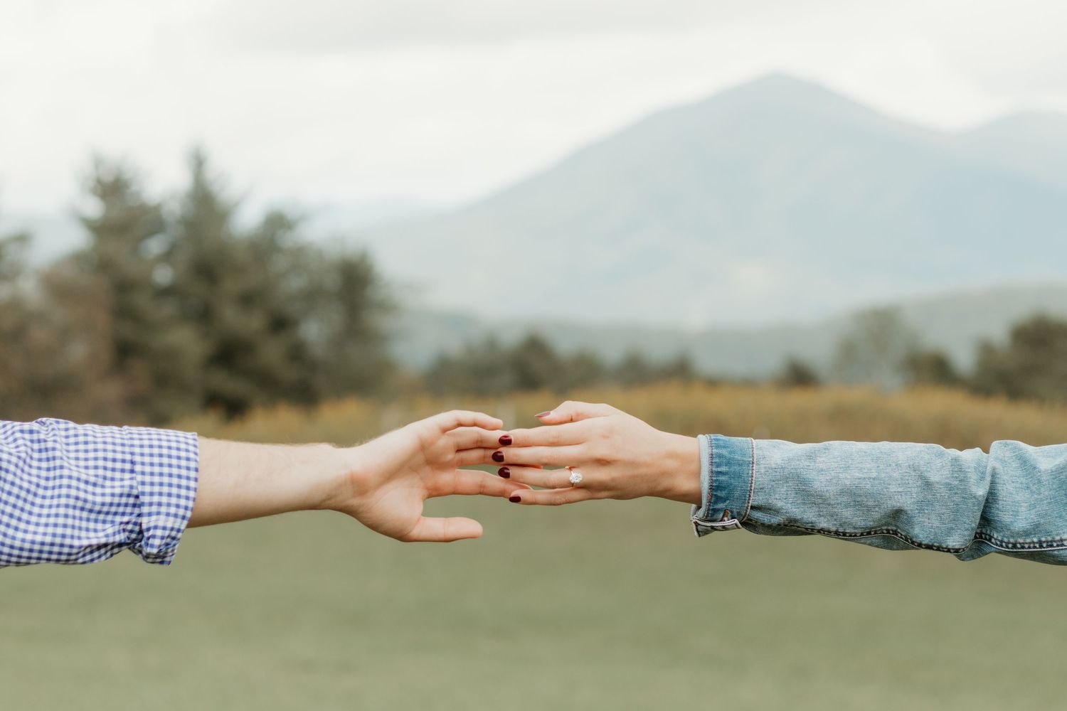 Two hands gently touching fingertips outdoors with mountains and green fields in the background.