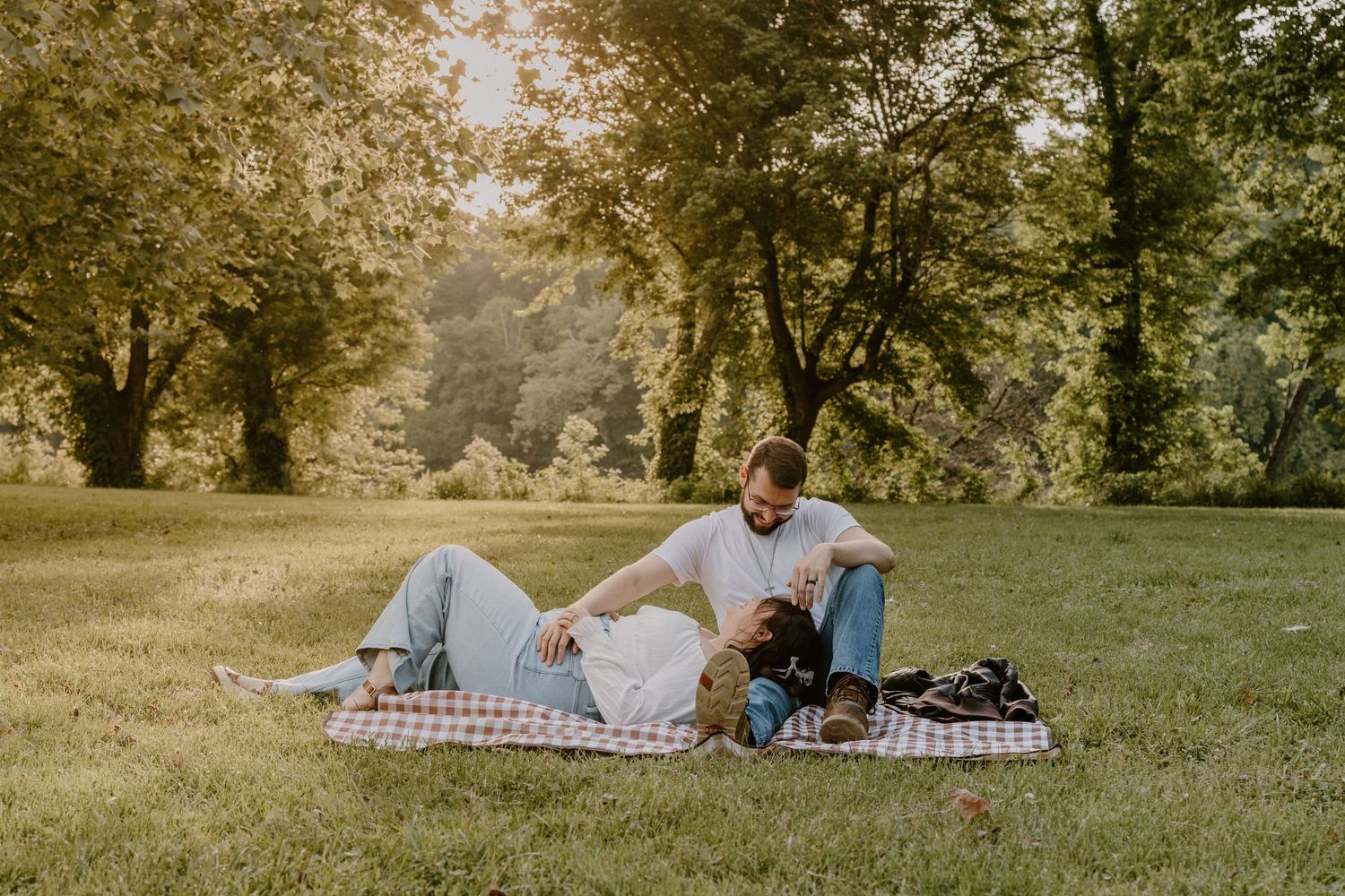 Couple relaxing on a checkered blanket in a sunny green park, surrounded by lush trees during a romantic outdoor picnic.