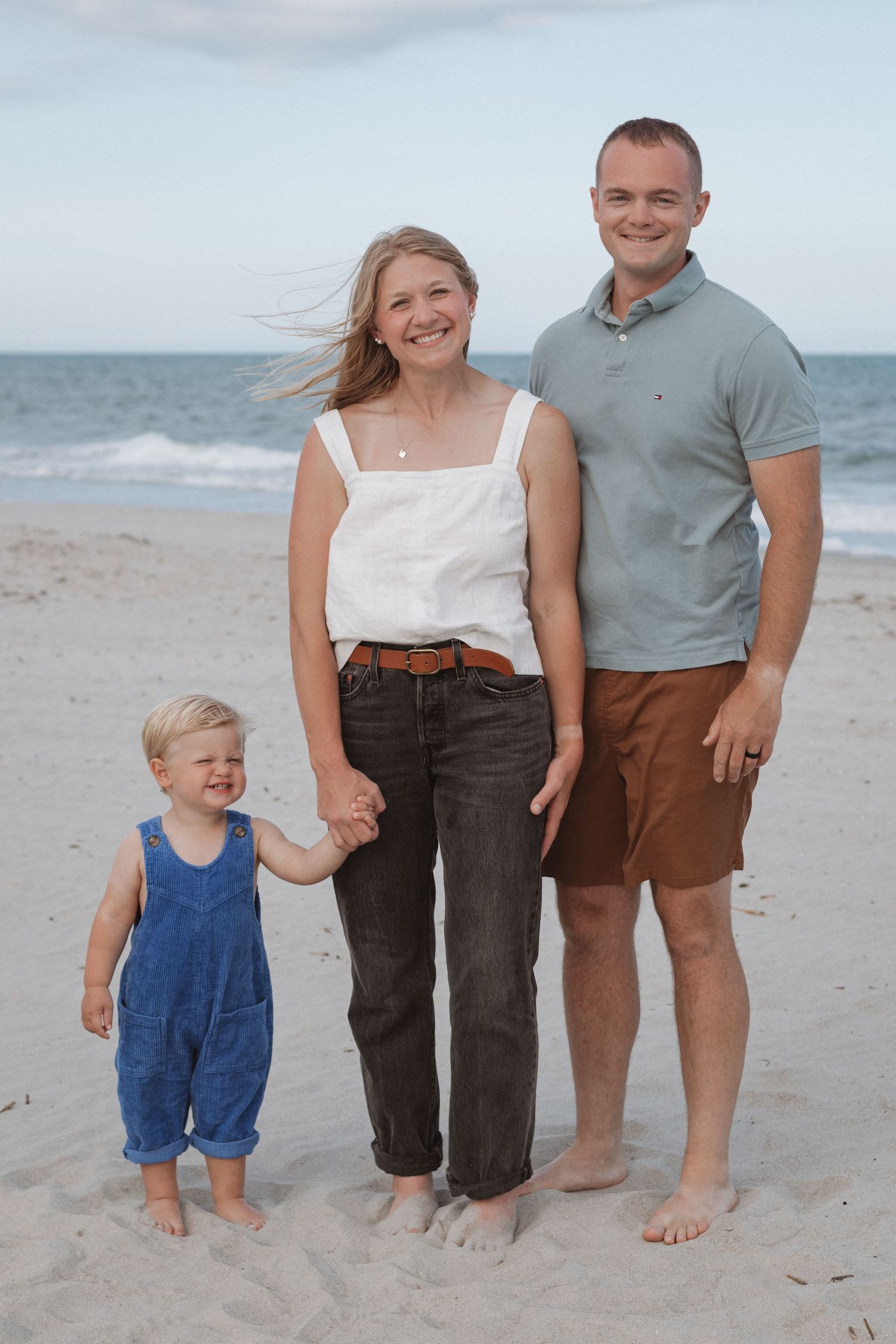 A family stands together barefoot on a sunny beach with ocean waves in the background.