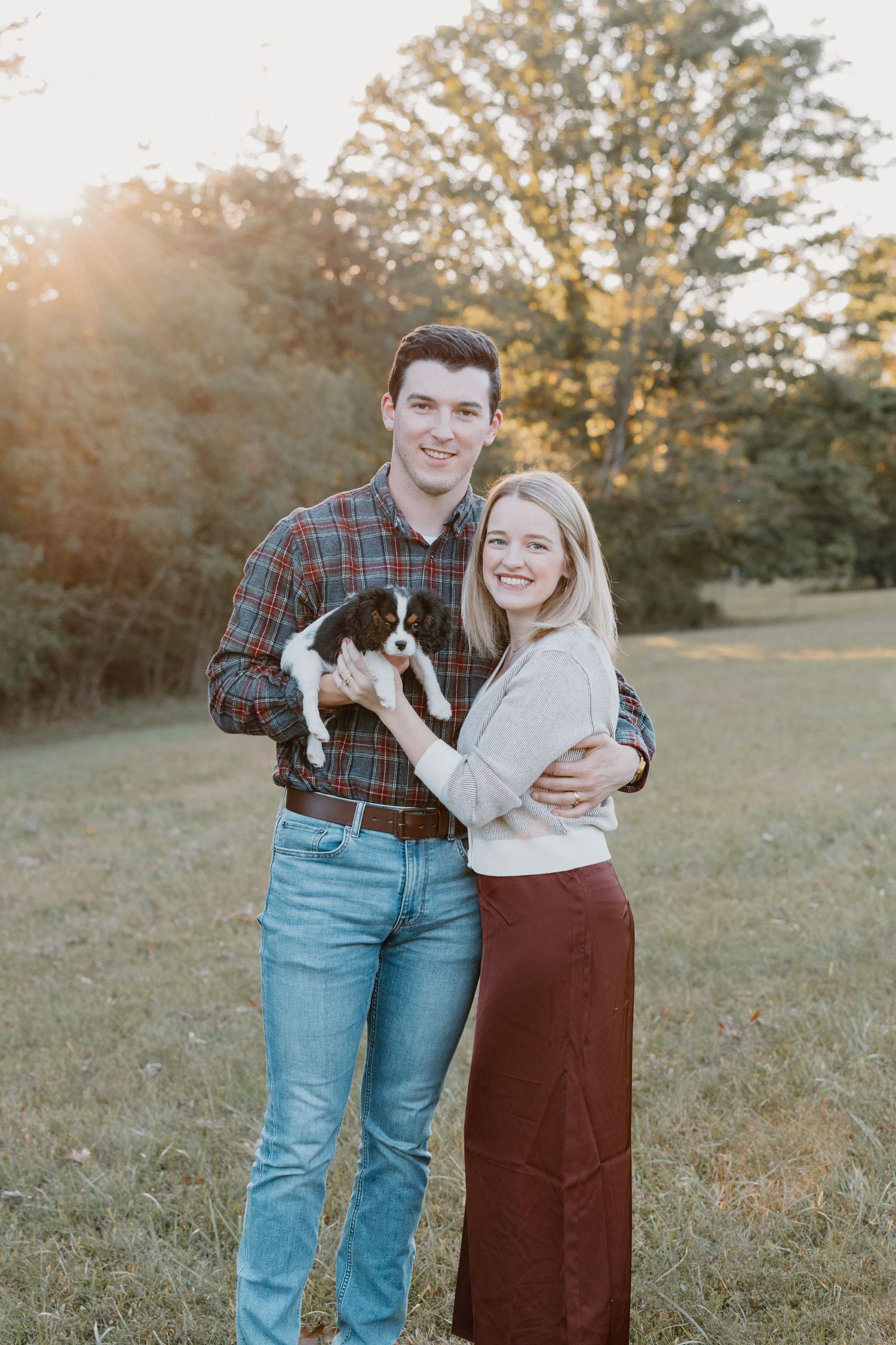 A couple stands together in a field at sunset while holding a small black and white puppy.