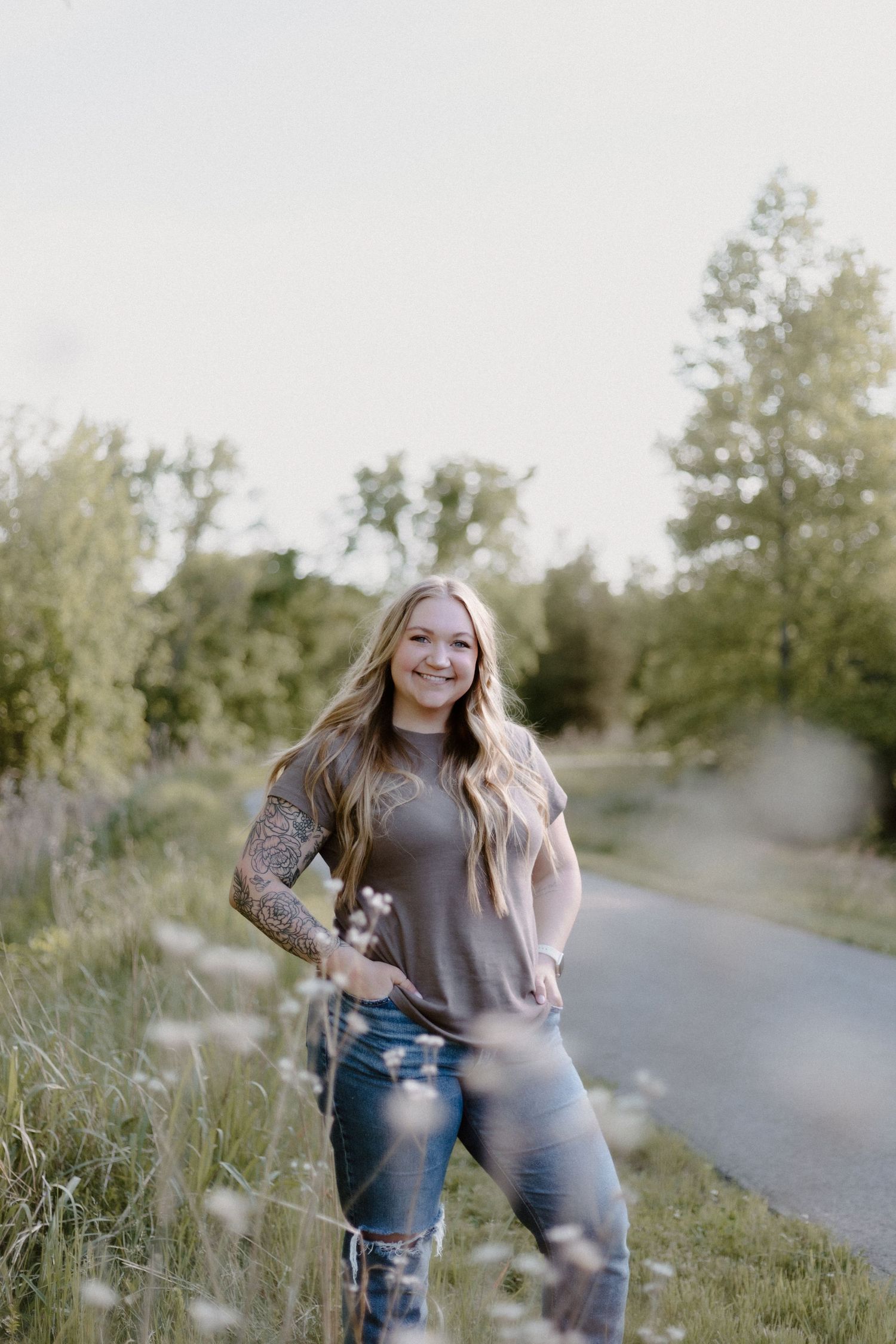 Young woman with long blonde hair and tattoo sleeve smiles on a scenic path surrounded by wildflowers.