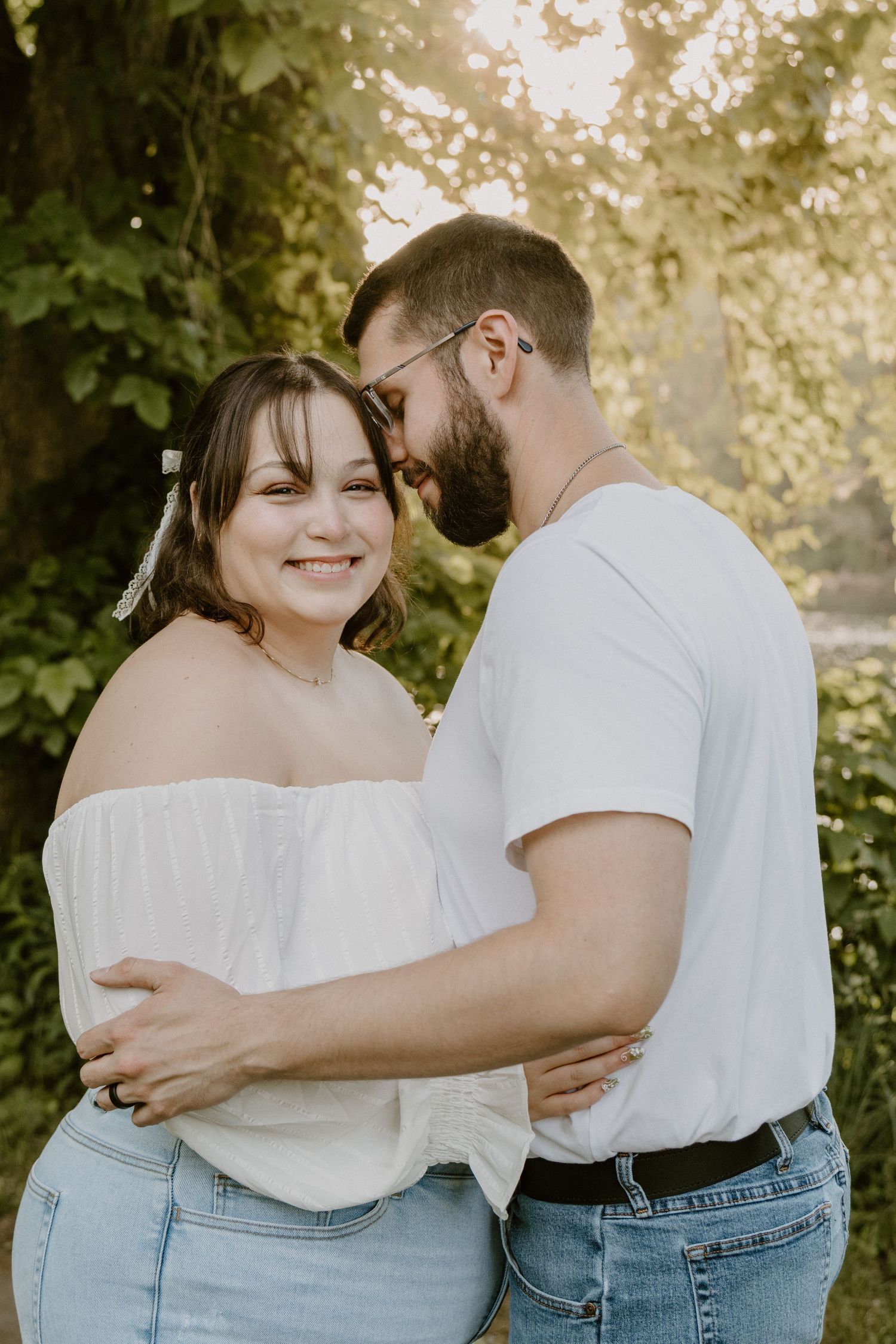 A smiling couple embraces outdoors in golden sunlight, woman in off-shoulder white top and man in white tee with jeans.