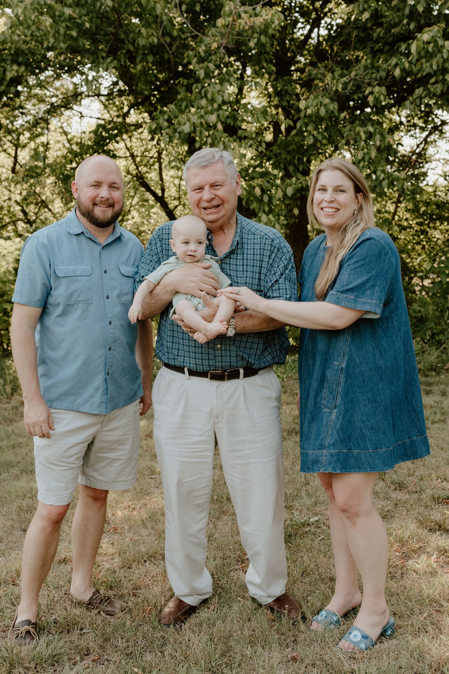 Three generations of family pose outdoors, with grandfather holding a baby, flanked by a young man and woman.