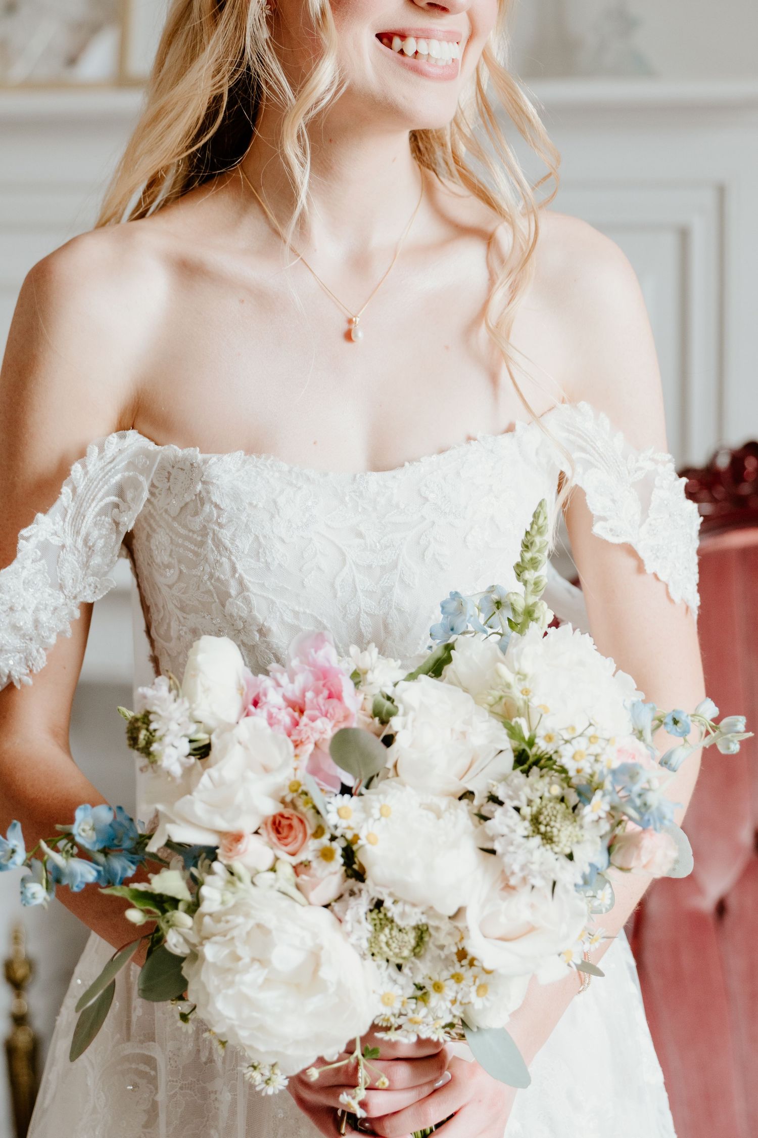 Smiling bride in off-shoulder lace wedding dress holding a pastel bouquet of white peonies and blue flowers.